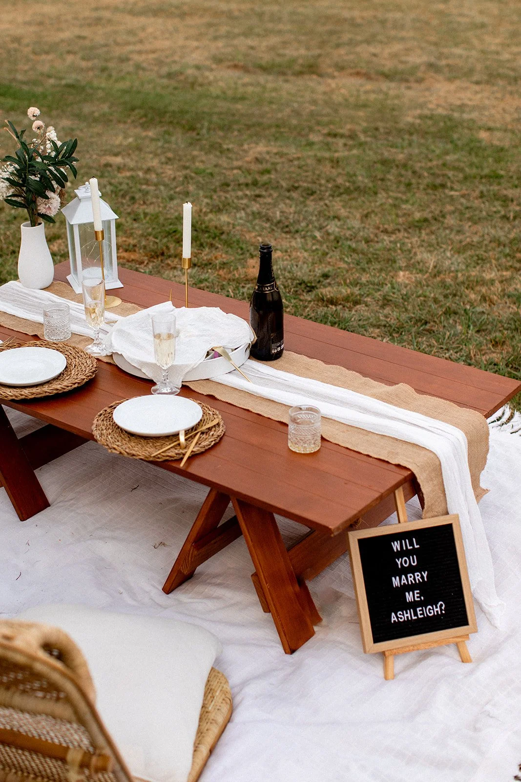 Outdoor dining setup on a wooden table with white plates, glasses, candles, a bottle of champagne, and decorative elements like a paper lantern and a floral arrangement; a blackboard sign asking 'Will you marry me, Ashleigh?'