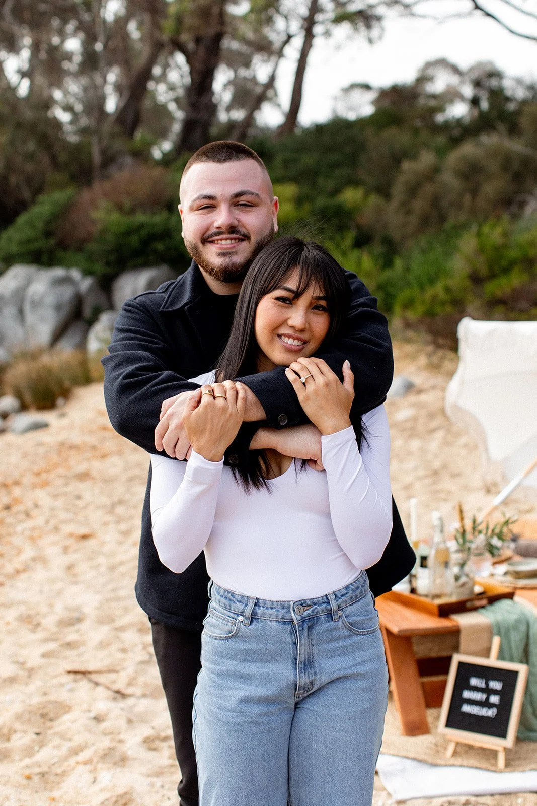 A couple hugging on a sandy beach with green trees in the background, with a table of snacks and bottles in the distance.