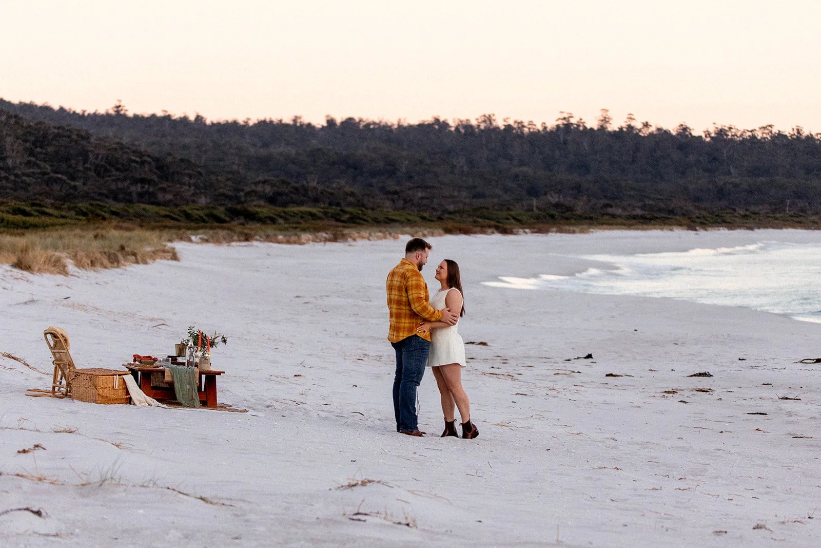 A couple on a beach during sunset, standing close and smiling at each other with a picnic setup nearby.