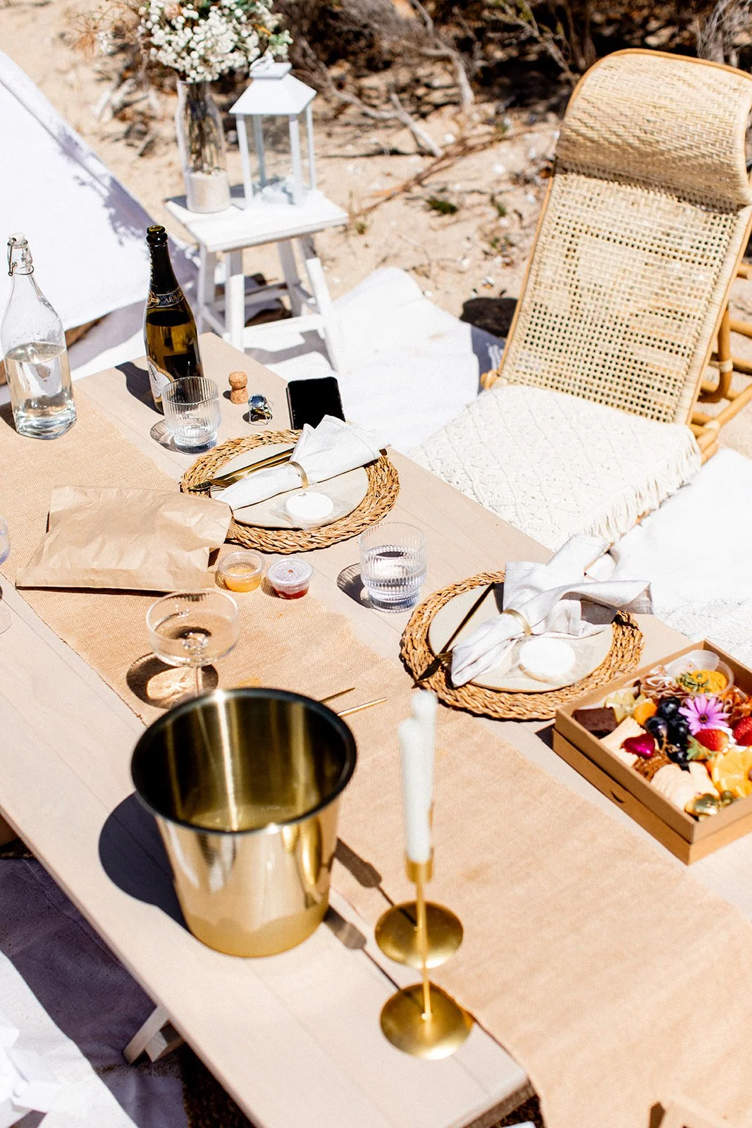 Outdoor table set for a celebration with plates, napkins, glasses, a bottle of champagne, a cheese and fruit platter, and a golden candle holder with a candle. The setting is on a sandy surface with a woven chair nearby, surrounded by decorative item