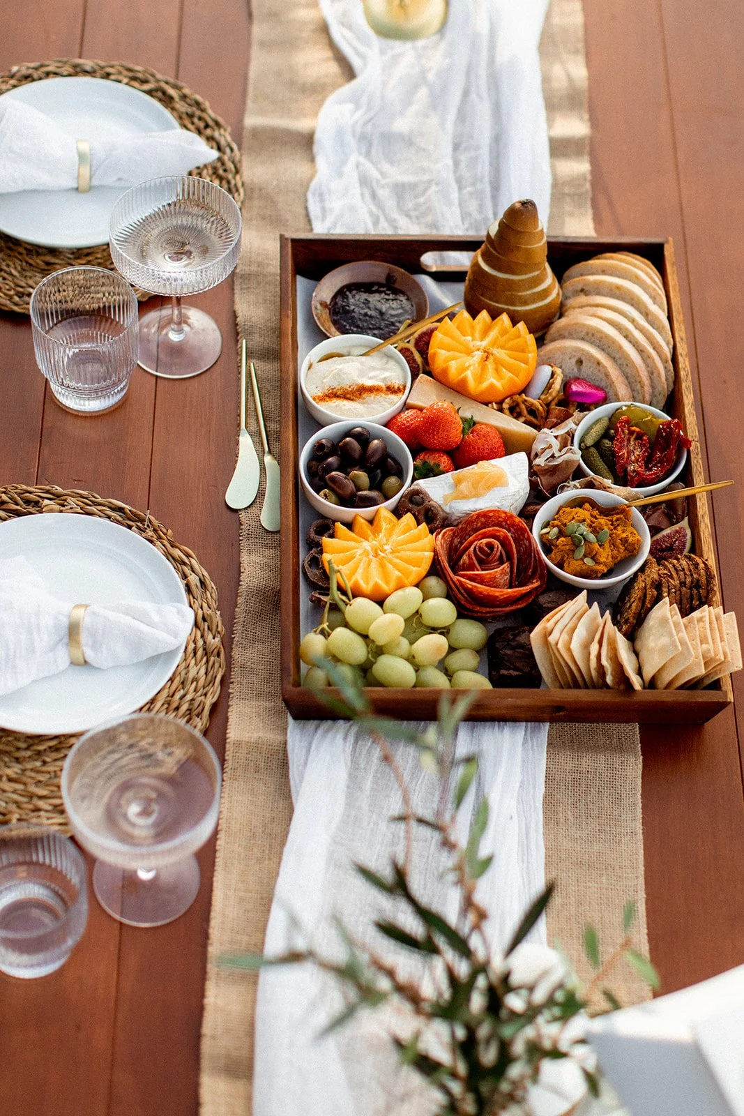 A charcuterie board with sliced bread, cheeses, strawberries, grapes, olives, figs, dips, and spreads on a dining table set with plates, glasses, and cutlery.