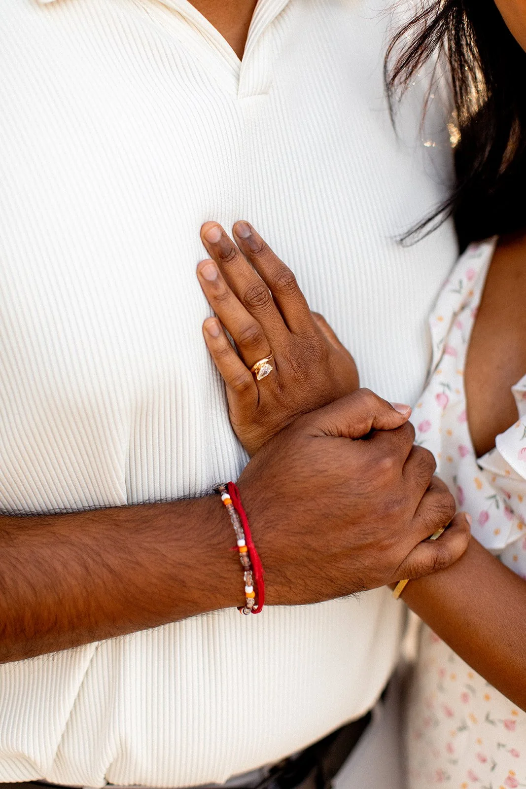 A close-up of a couple's hands showing a wedding ring, with the woman's hand resting on the man's chest, both wearing simple jewelry, including a red and white beaded bracelet.