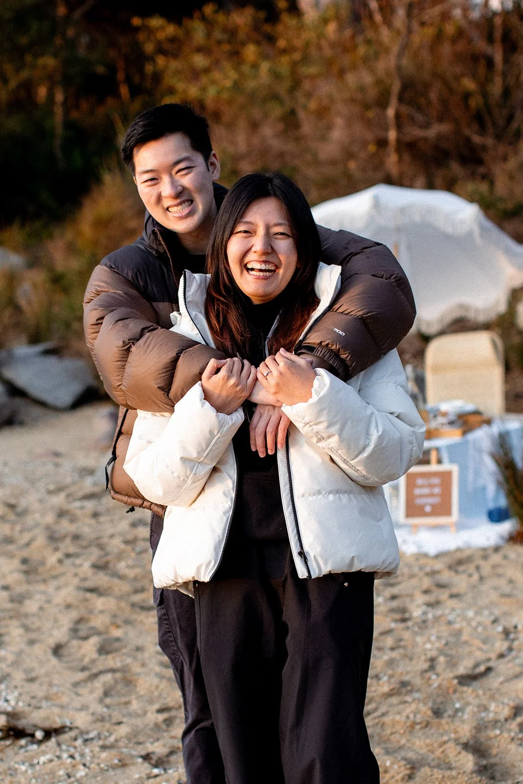 Two people smiling and hugging outdoors during daytime, with trees and a table in the background.