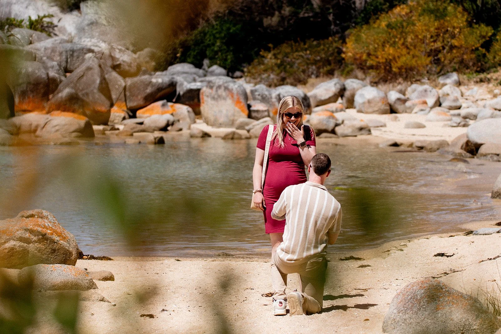 A man kneeling on one knee in front of a woman in a red dress, who appears to be surprised and happy, at the edge of a river with rocks and autumn foliage in the background.