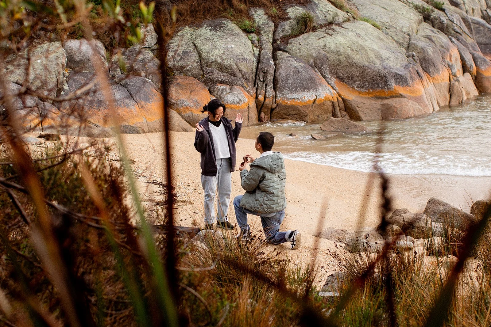 A man on one knee proposing to a woman on a beach, with rocky cliffs in the background.