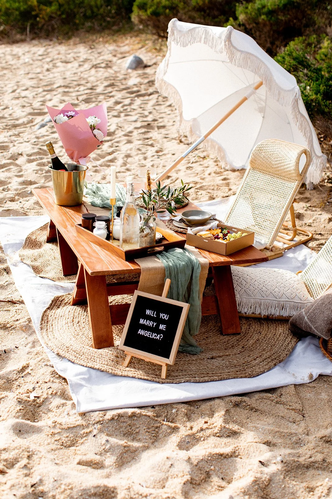 A romantic beach picnic setup with a low wooden table on sand, decorated with flowers, drinks, and snacks. There are two woven chairs and a white parasol for shade. A small chalkboard sign reads, 'Will you marry me Angelica?'