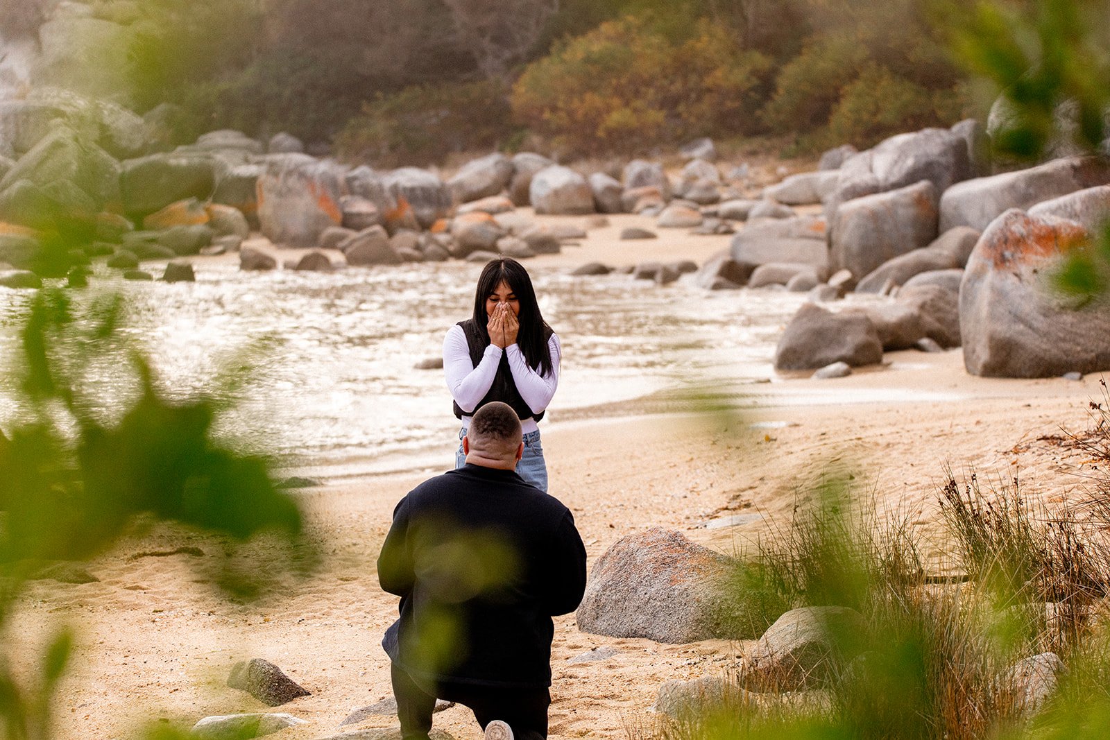 A young woman appears emotional, holding her hands to her face while standing in front of a man kneeling on a sandy beach near rocks and greenery with a river and trees in the background.
