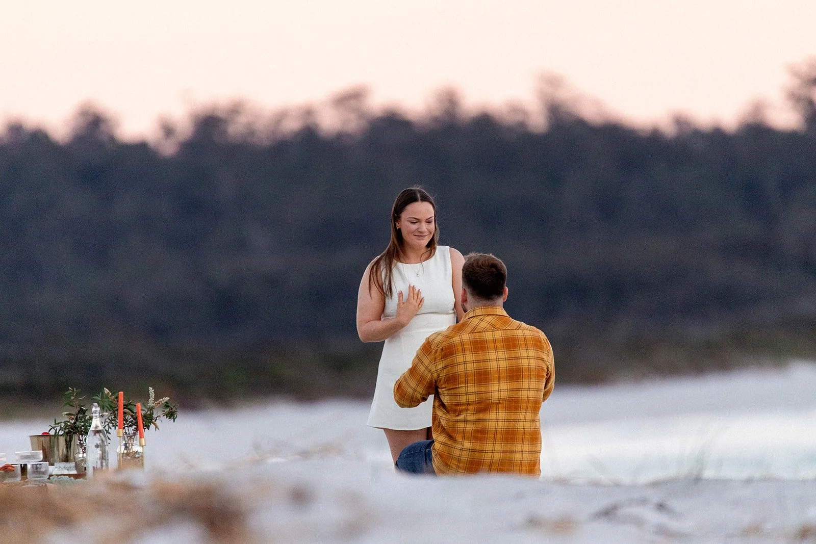 A man proposing to a woman on a beach at sunset, with a picnic setup nearby.