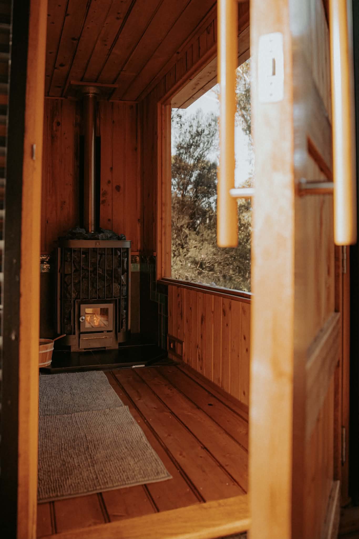 A cozy wooden room featuring a wood-burning stove, wood-paneled walls and ceiling, a small corner window overlooking trees, a rectangular area rug on the floor, and a basket near the stove.
