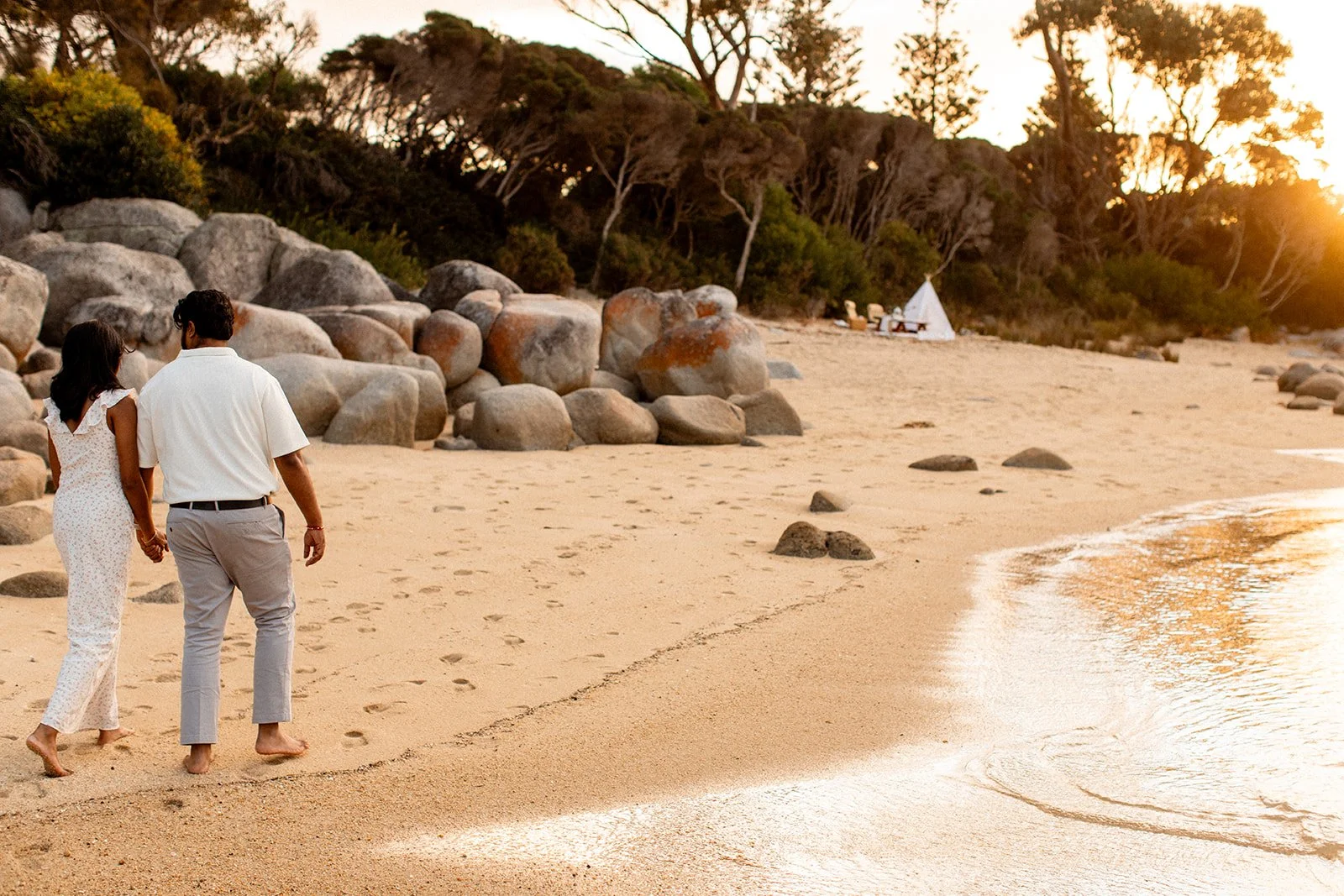 A couple holding hands walks barefoot on a sandy beach during sunset, with large rocks and trees in the background.