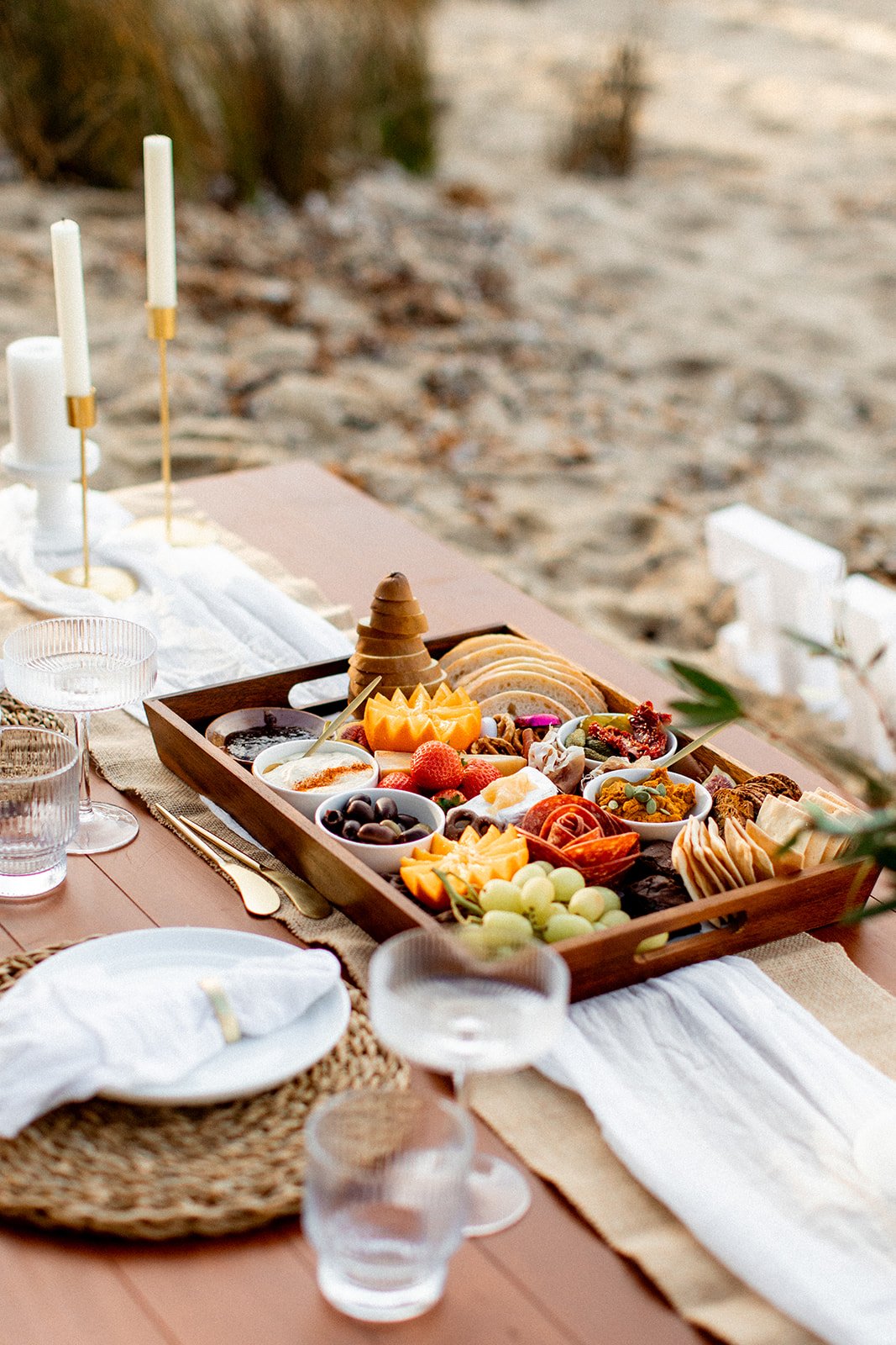 A picnic table set with a wooden tray of assorted cheeses, fruits, and snacks, surrounded by glasses of water, candles, and a white tablecloth on a sandy beach.