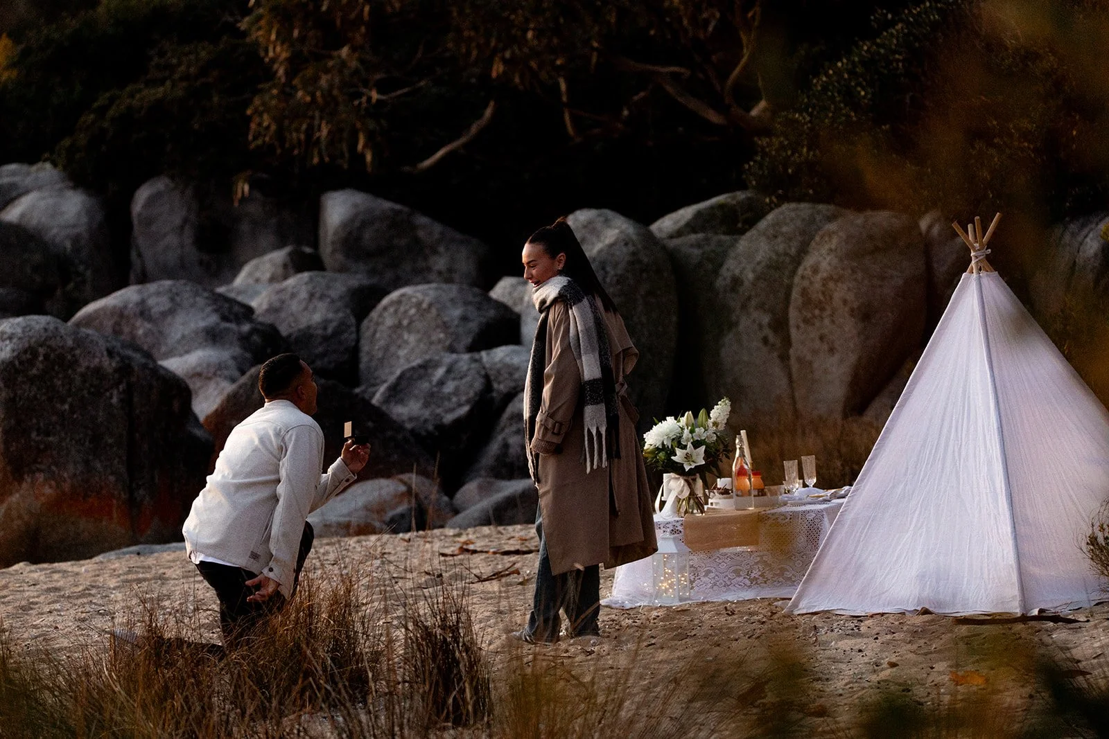 A woman proposing to a woman outdoors near a small teepee with a table set for a celebration, surrounded by rocks and trees at dusk.