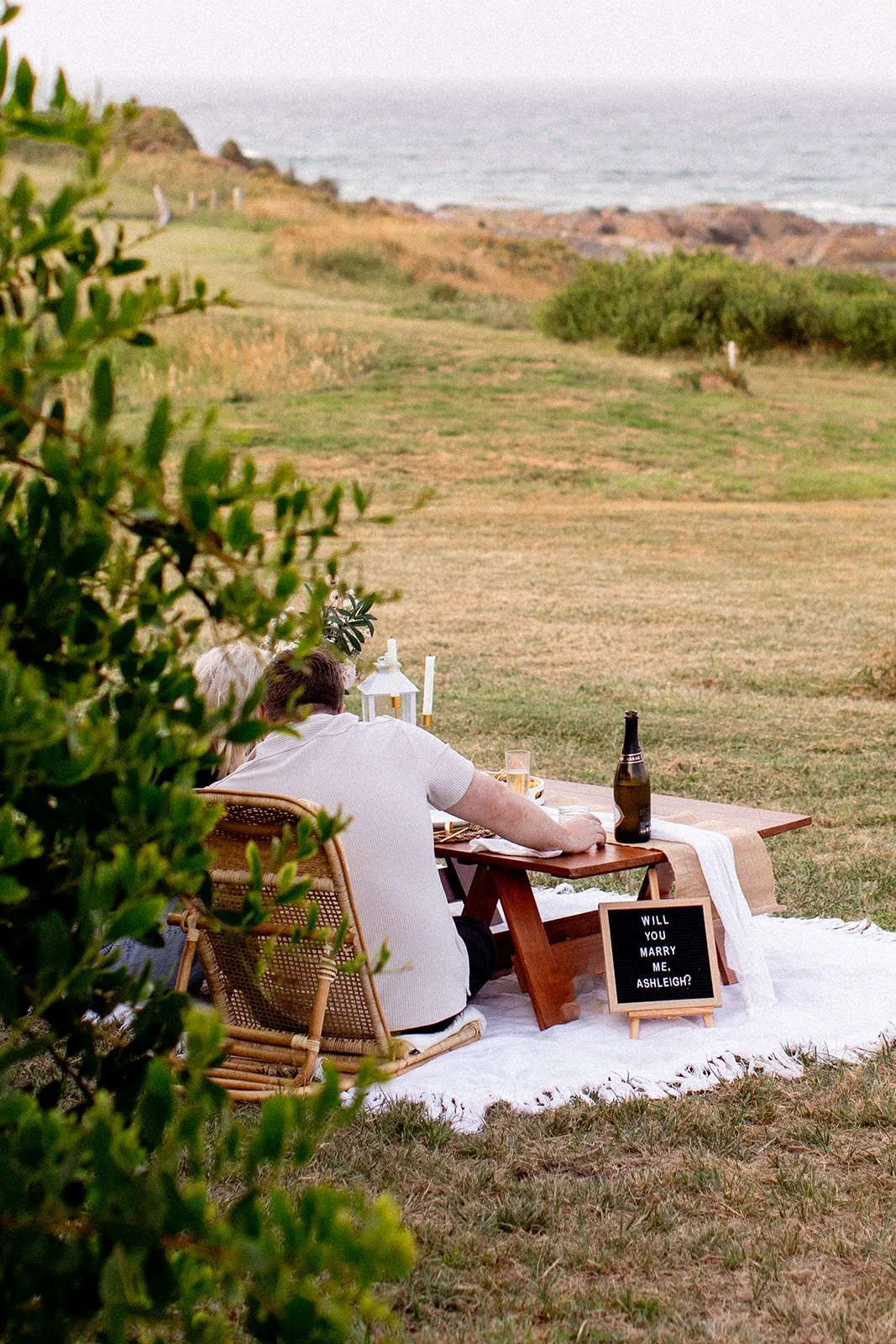 A couple seated on a picnic blanket enjoying a romantic proposal on a grassy outdoor area near the coast, with a small blackboard sign asking "Will you marry me, Ashleigh?" surrounded by drinks, lanterns, and candles.