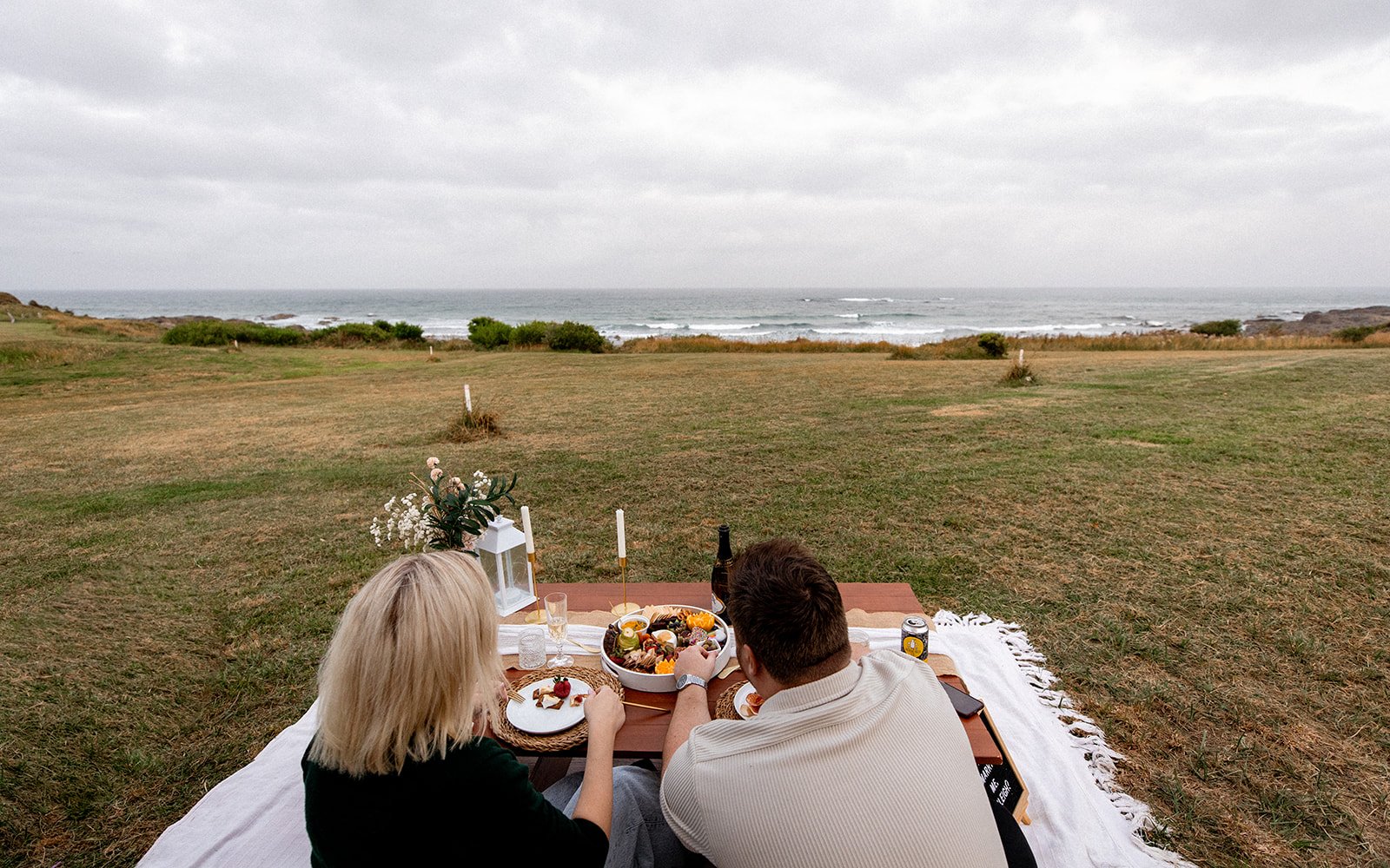 A couple having a picnic on a blanket at the beach, with food and drinks on a low table, overlooking the ocean under cloudy skies.