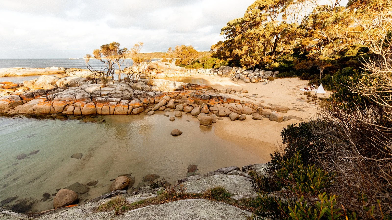 Coastal scene with large rock formations, sandy beach, and trees. A white tent is visible on the beach, with some people nearby.