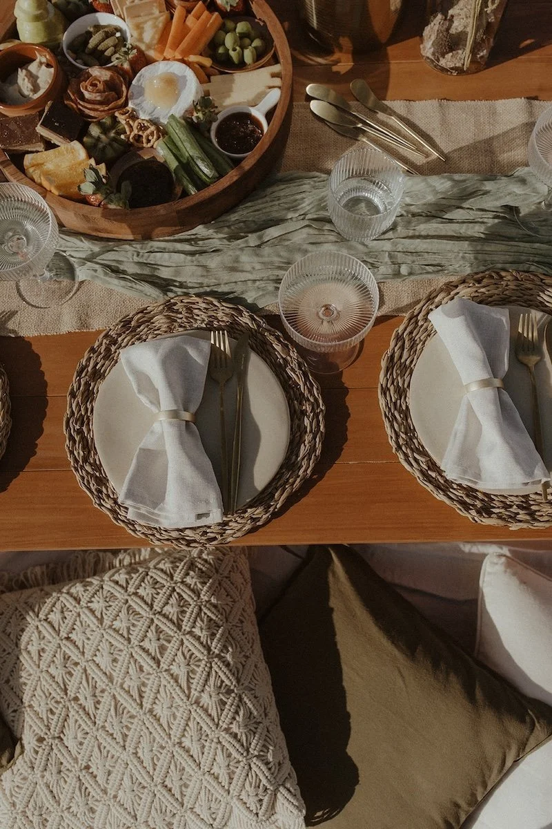 A dining table set with three round plates, each with a white cloth napkin tied with a gold band, silverware, and clear textured glasses. A central wooden serving tray contains various cheeses, vegetables, dips, and condiments, with a beige table runner underneath.