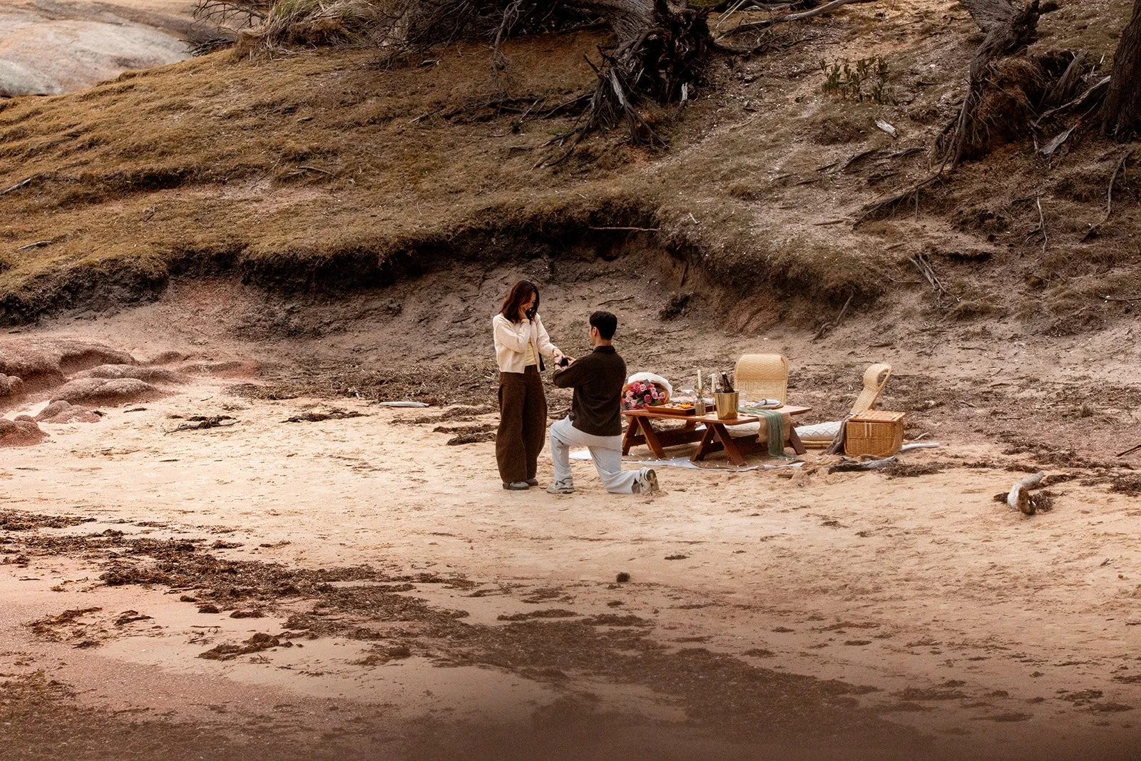 A couple is having a wedding ceremony on a sandy beach with a backdrop of a rocky, grassy hillside. The man is kneeling and placing a ring on the woman's finger. There is a small table with flowers, wine glasses, and other items next to them.