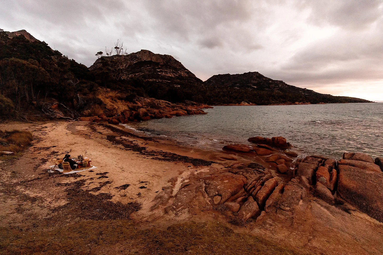 A person relaxing on a beach during dusk with two chairs and a small table, surrounded by rocks and trees, overlooking the ocean with hills in the background and a cloudy sky.