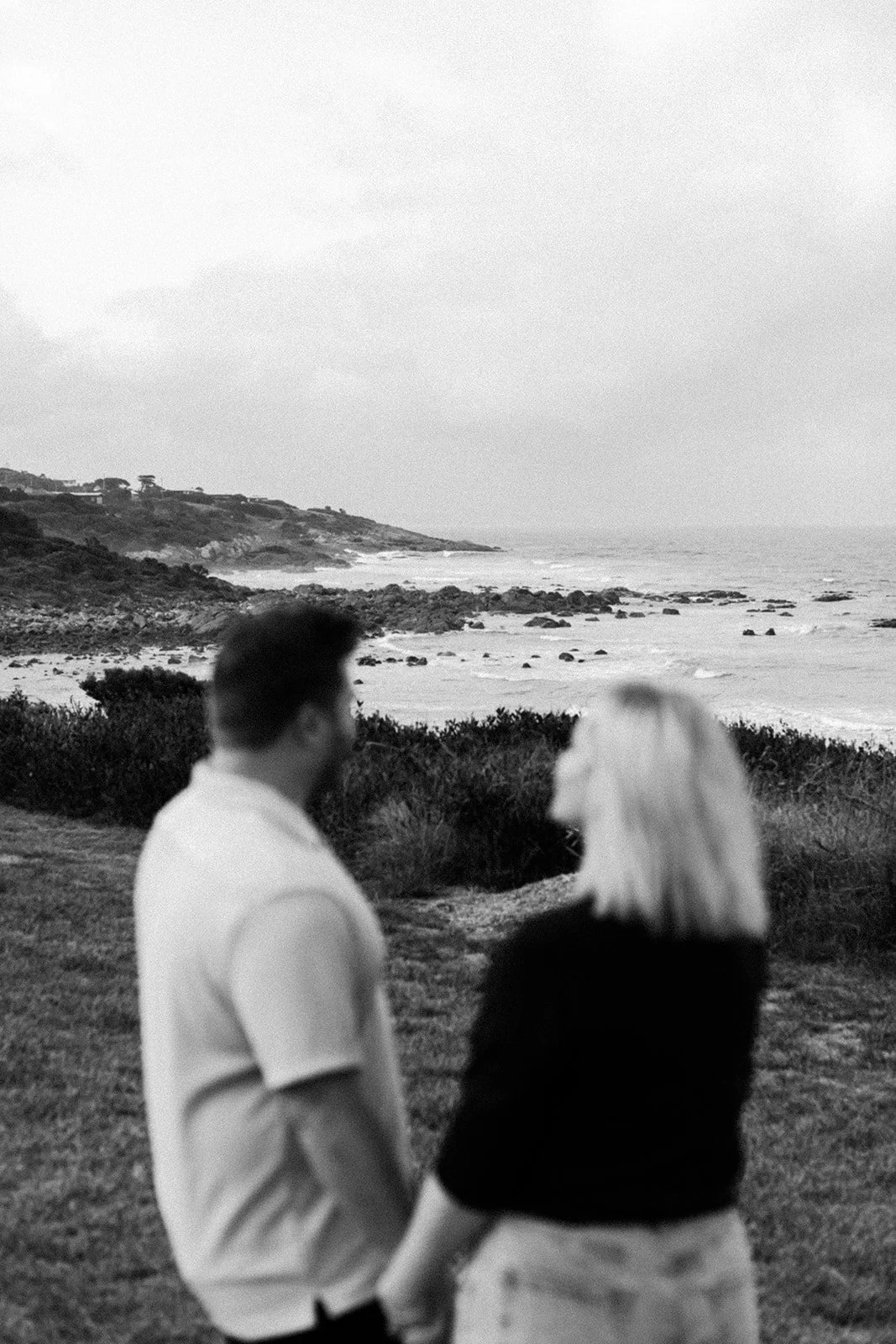Two people holding hands and talking on a grassy area by a rocky coastline with ocean waves, hills, and sky in the background.