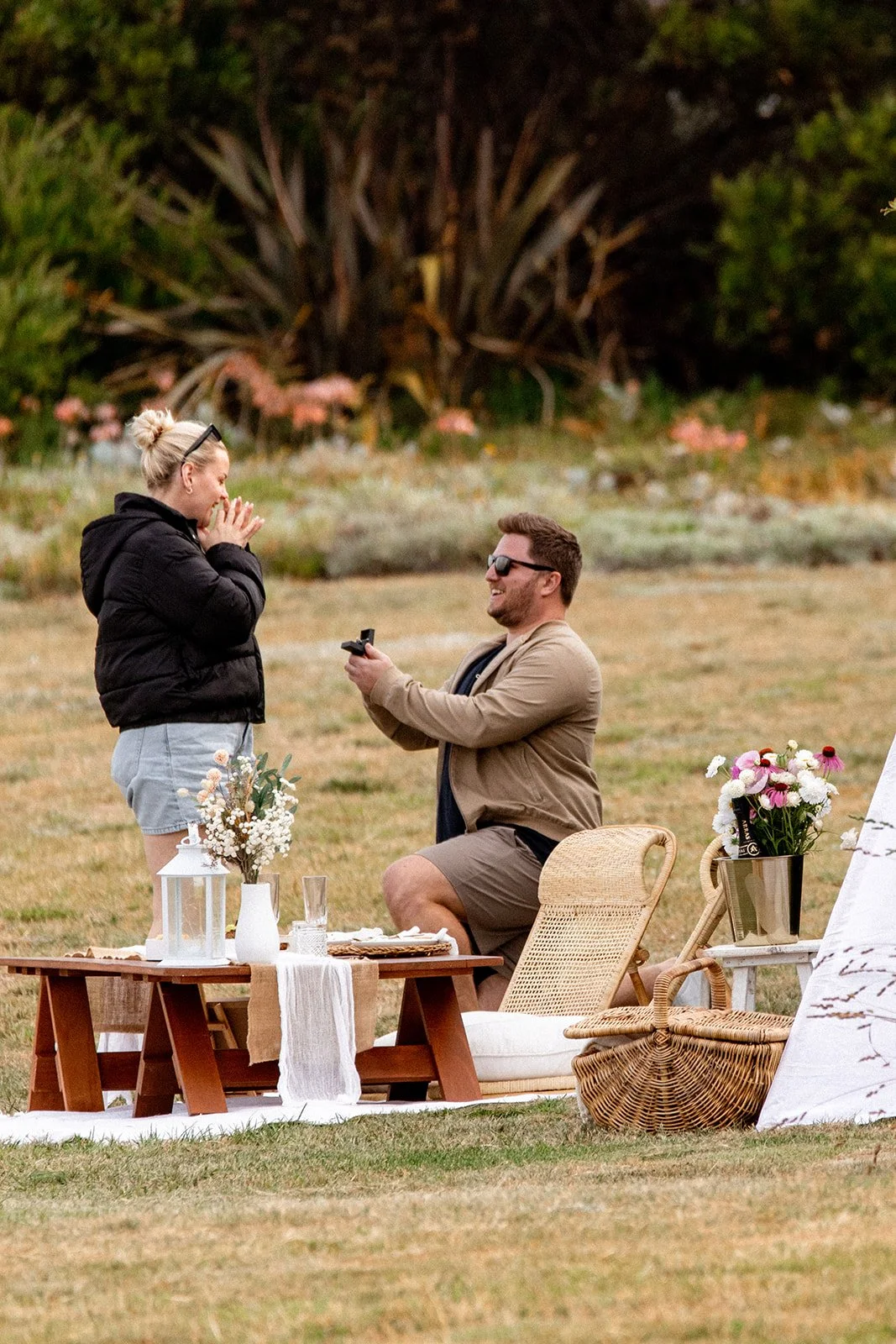 A man is proposing marriage to a woman outdoors during sunset, with the man kneeling and holding an open ring box, and the woman reacting happily. There are flowers and a picnic setup nearby.