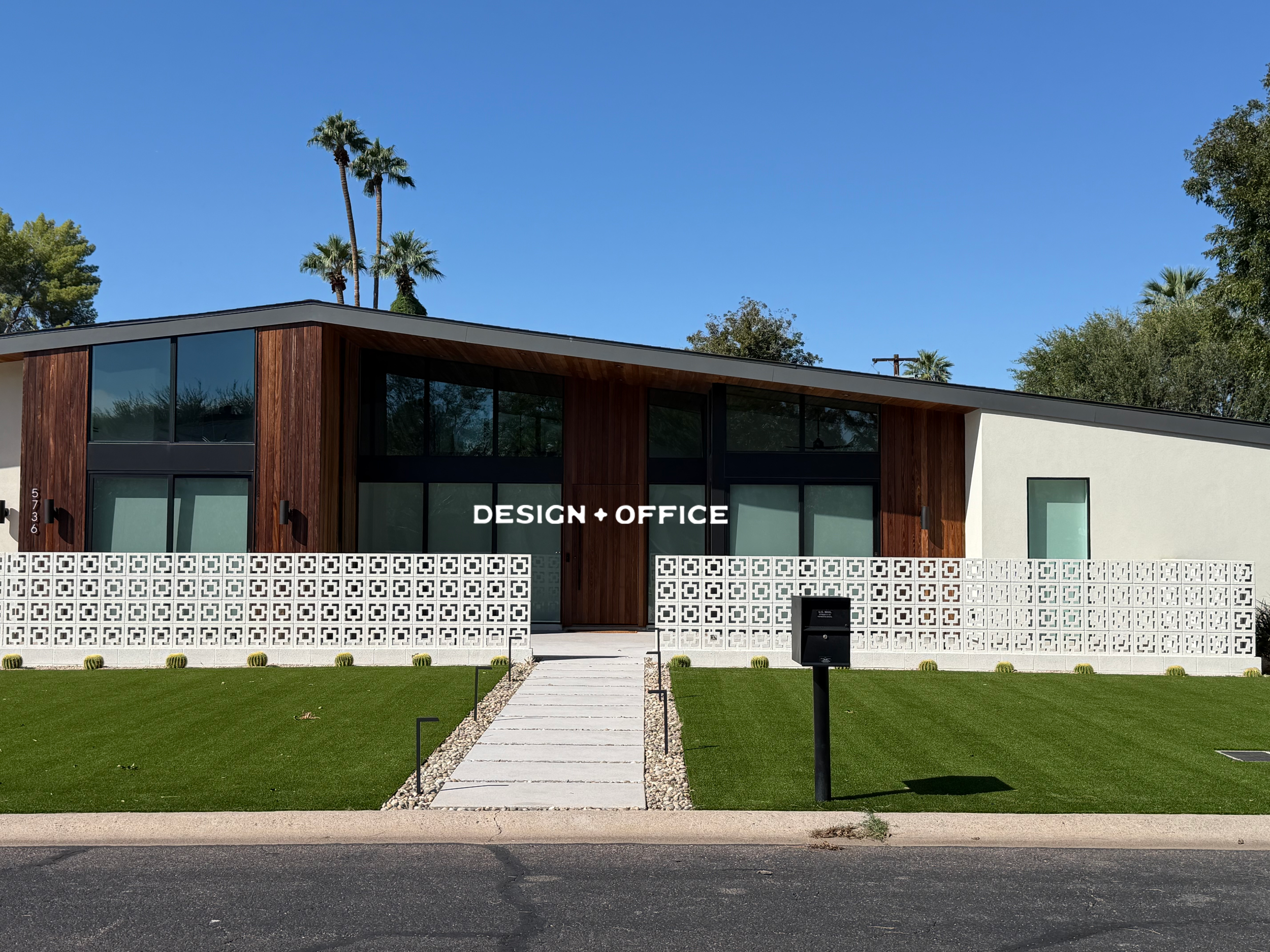 Modern two-story building with large glass windows, a wooden and white exterior, and a white decorative fence in front. There is a walkway leading to the entrance, surrounded by green grass, with palm trees and other trees in the background. The sky is clear and blue.