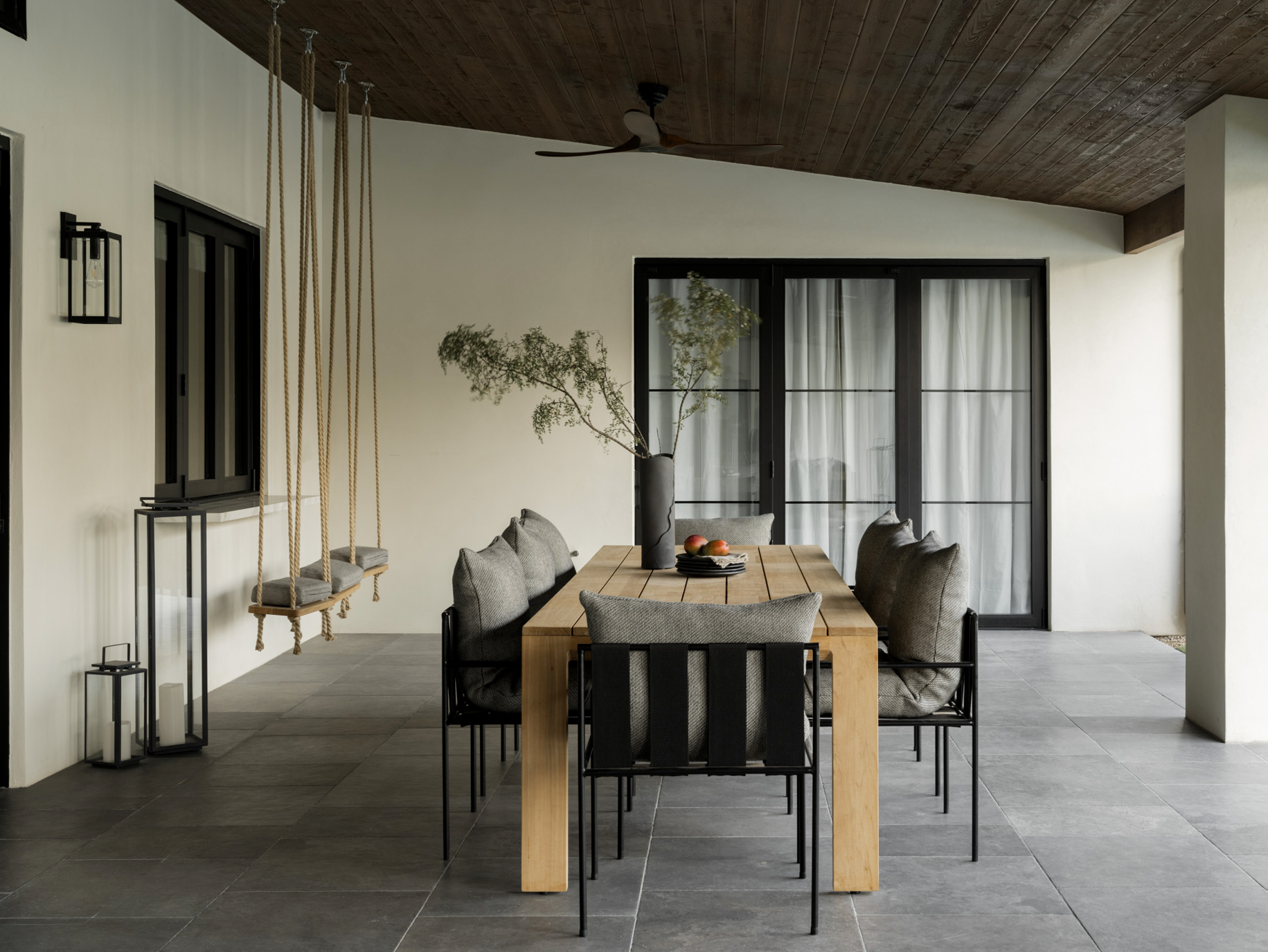 Modern dining room with a wooden table, six upholstered chairs, a black vase with greenery, and a black sliding glass door with white curtains. A ceiling fan and wooden ceiling are visible, along with hanging rope swings and decorative candle holders by the wall.