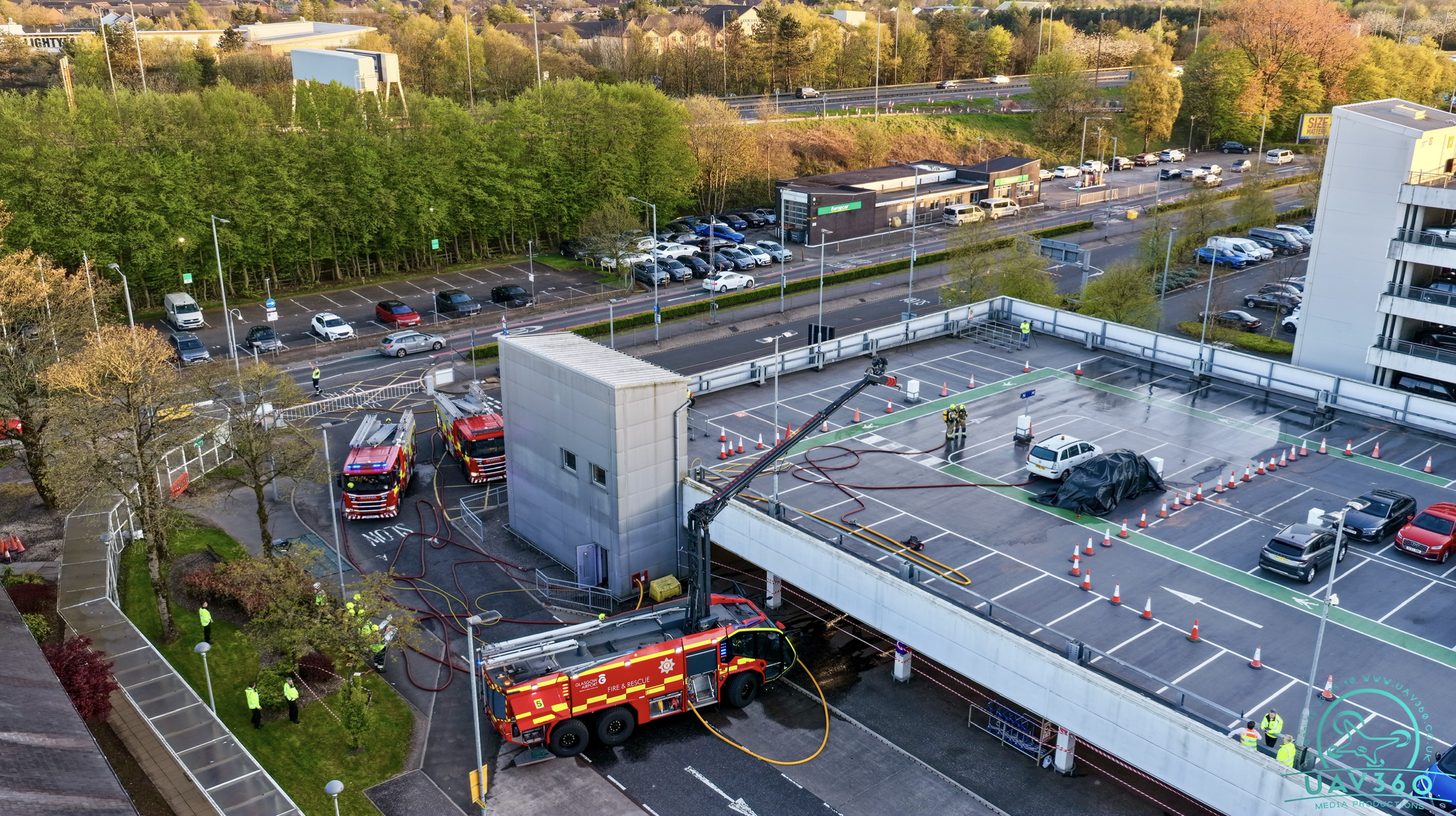 Aerial view of a parking lot near a building with several emergency vehicles and personnel present, some hoses on the ground, and a covered object, with a background of cars on a highway and trees.