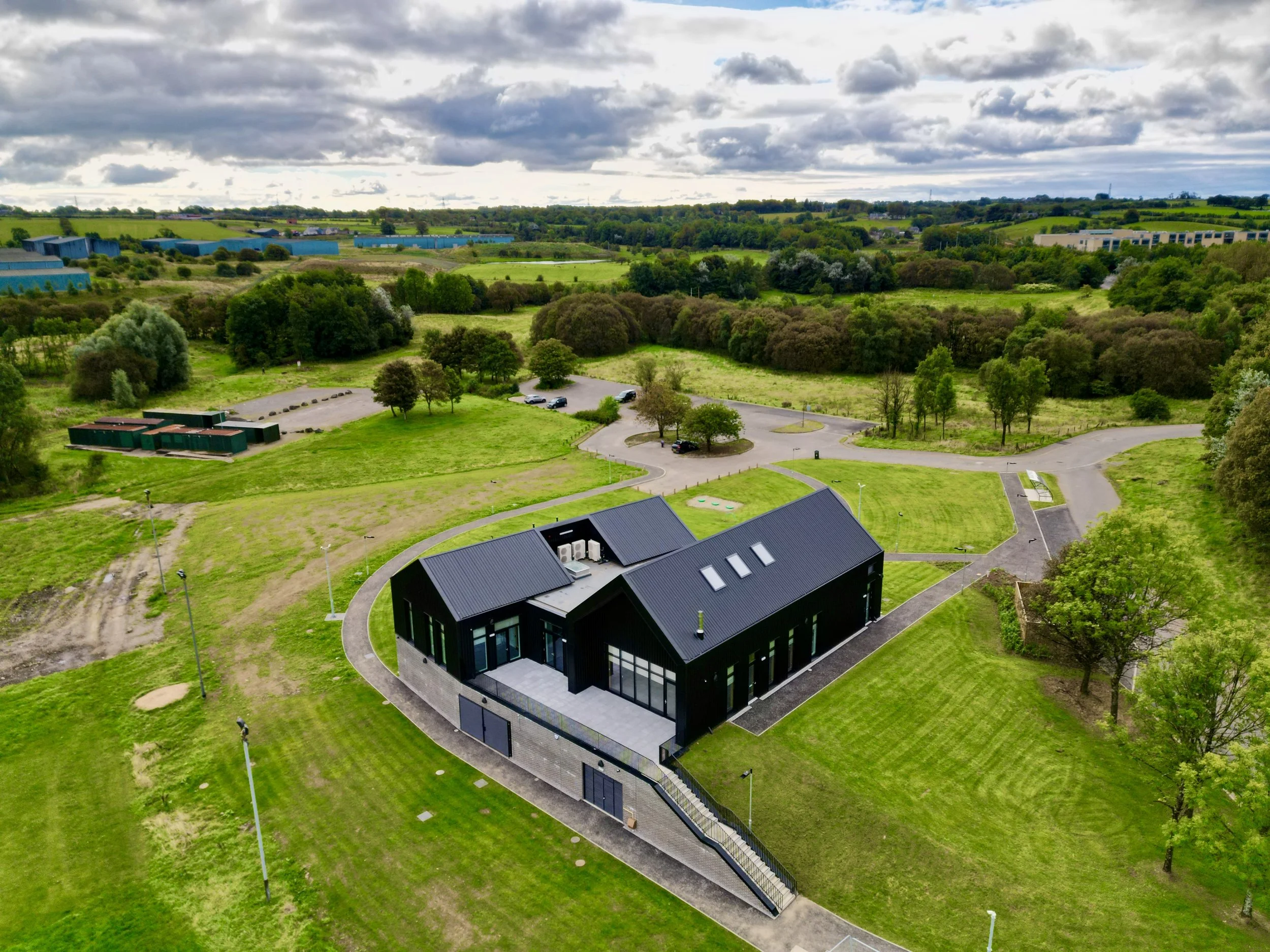 A modern black house with large glass windows surrounded by green grass and trees, located in a scenic rural area with fields, clouds, and a parking lot nearby.
