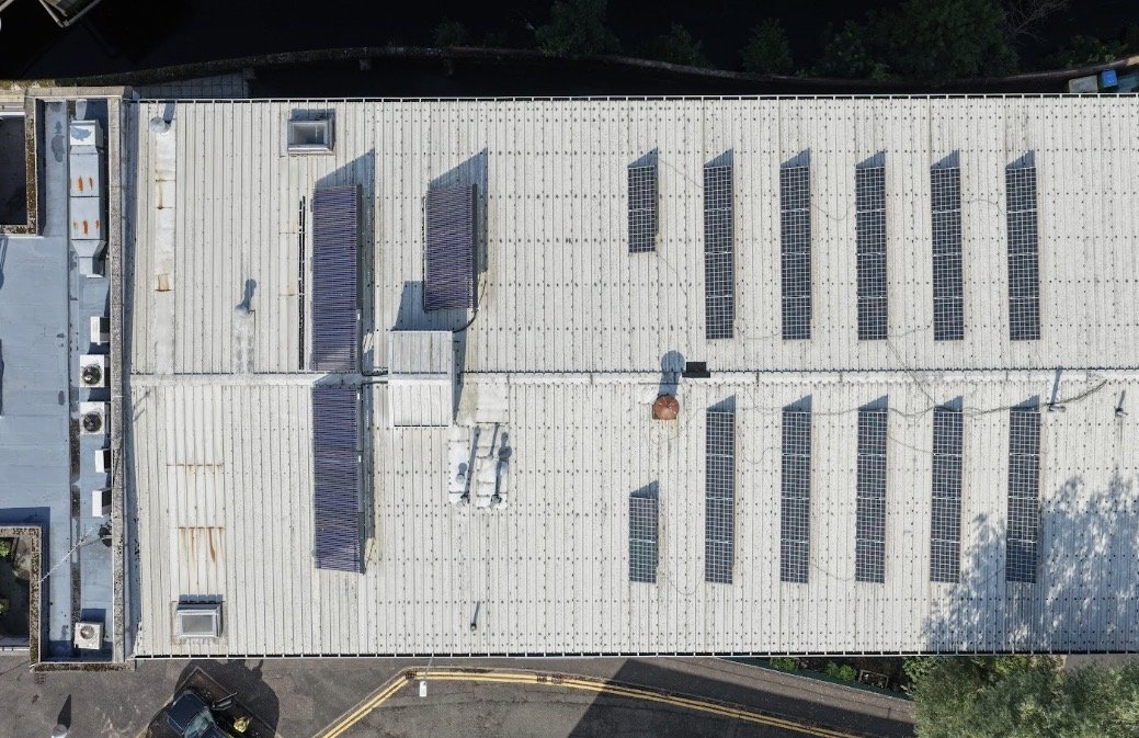 Large industrial fire with dark smoke billowing into the sky, flames visible at the base, near factory buildings and surrounded by a commercial or industrial area at dusk.