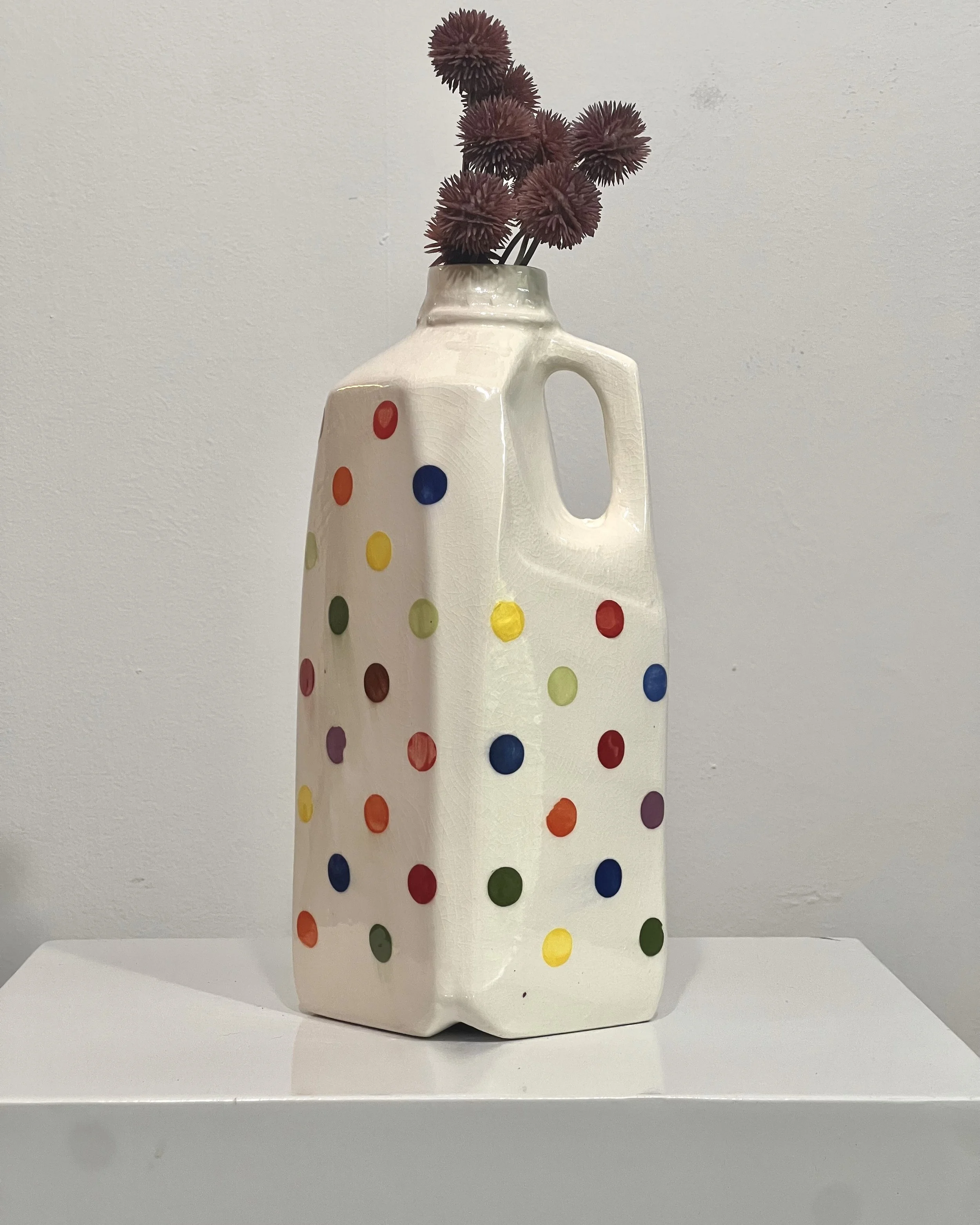 Tall white porcelain milk jug with hand-painted multicolored dots and integrated handle, displayed with burgundy pom-pom flowers.