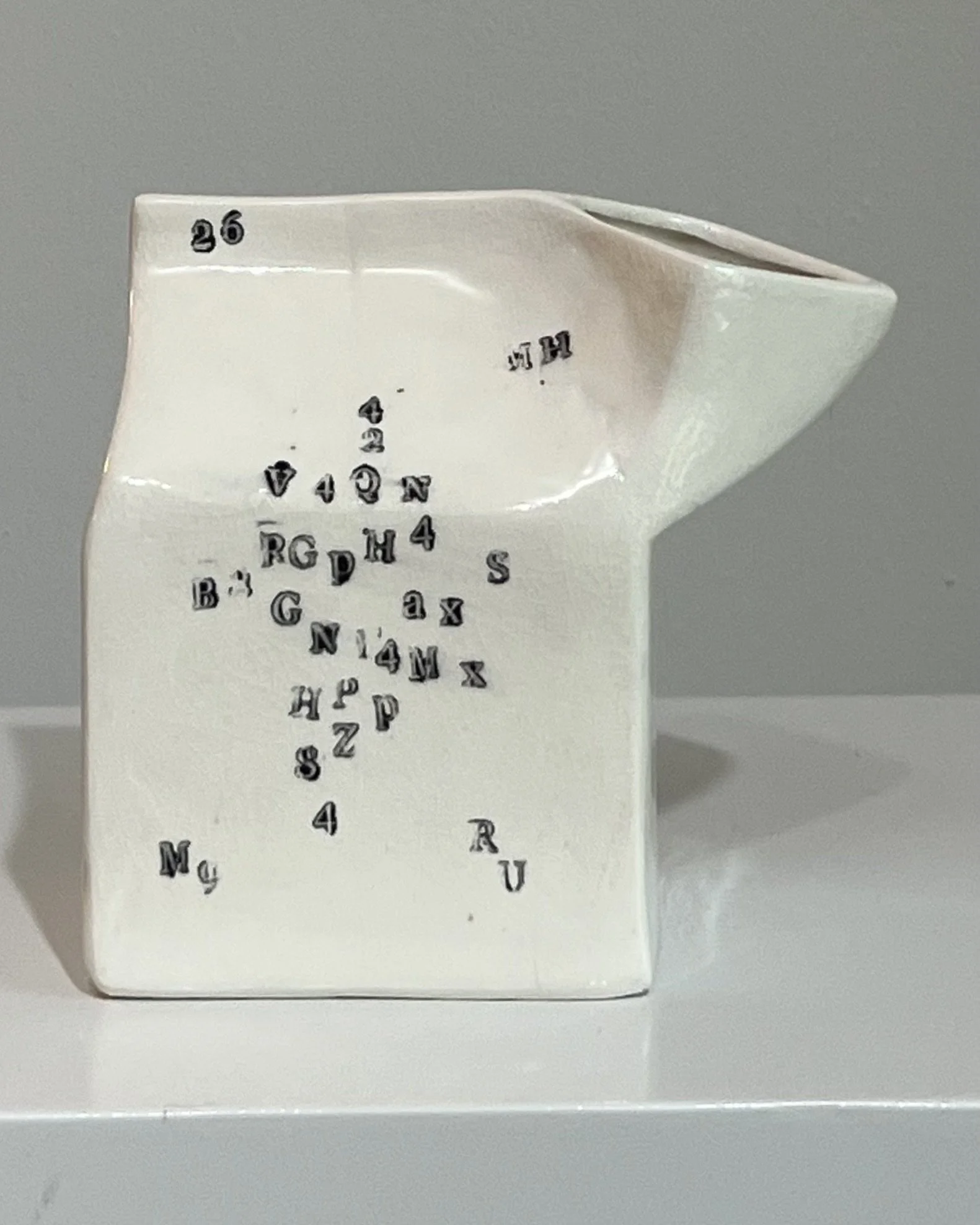 Front view of a handbuilt white porcelain milk carton with scattered black typographic impressions and a subtly compressed form.