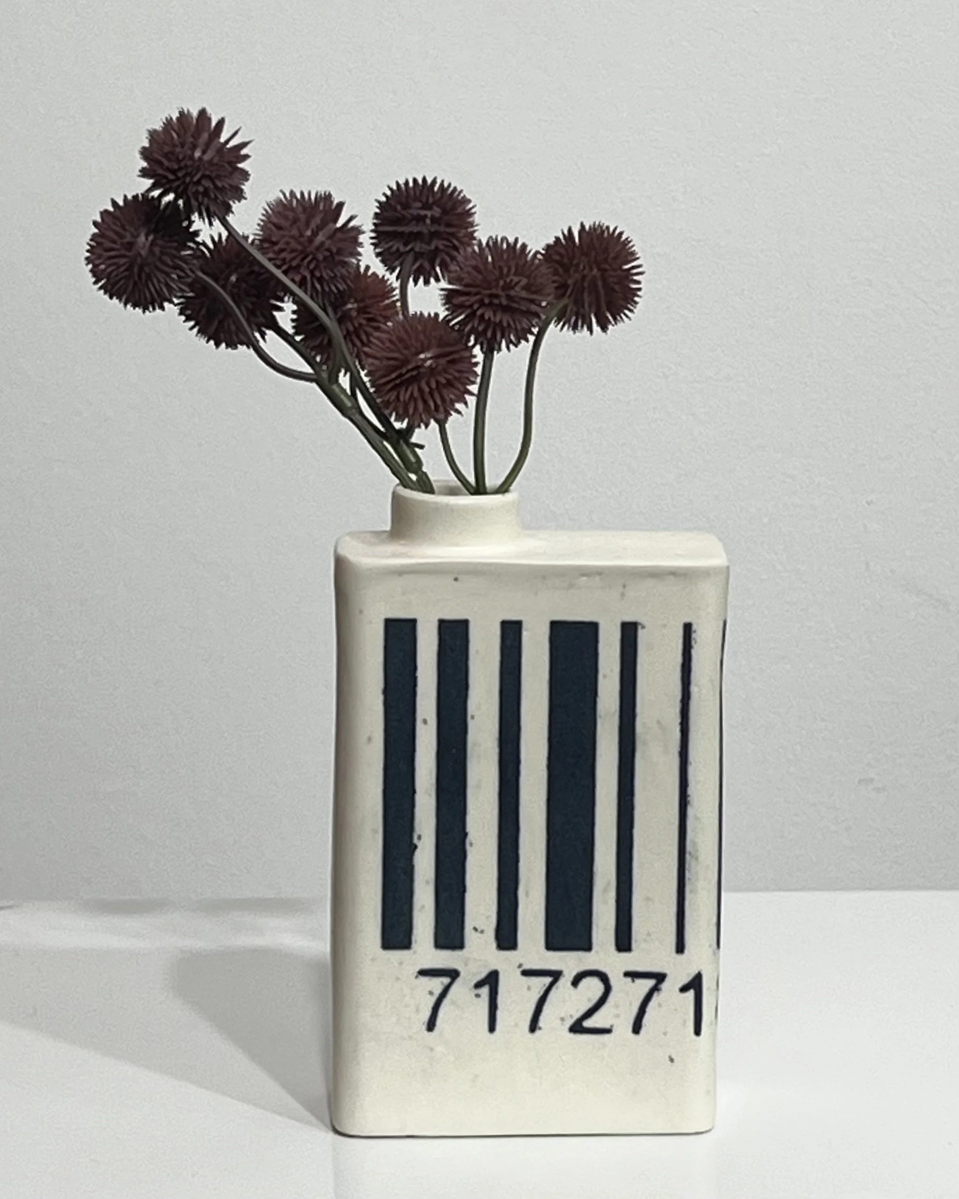 White porcelain milk carton vase with black barcode design and dark red dried flowers