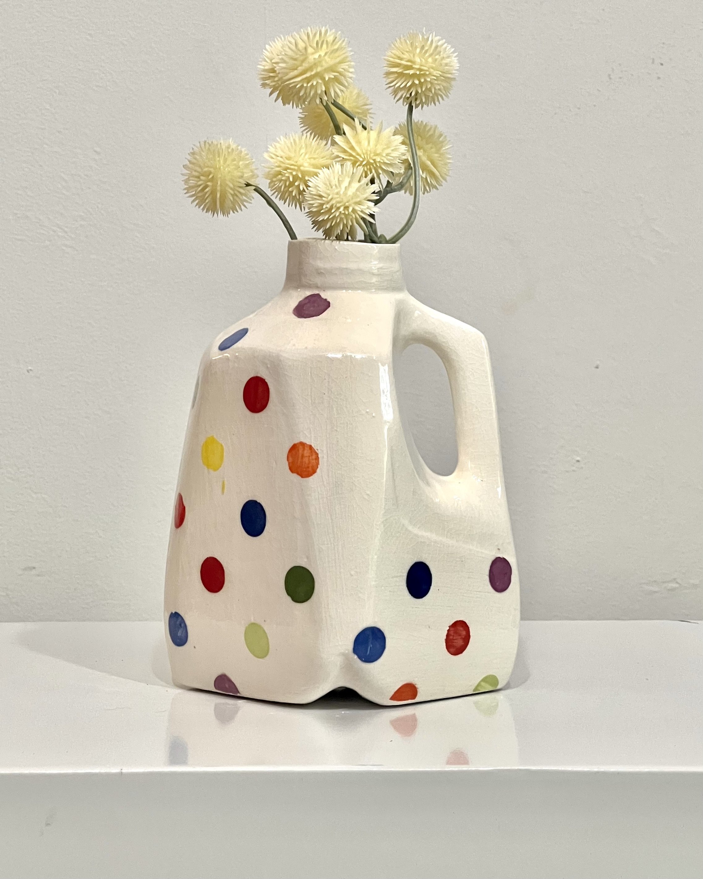 White porcelain milk jug with hand-painted multicolored dots and integrated handle, displayed with cream-colored flowers.