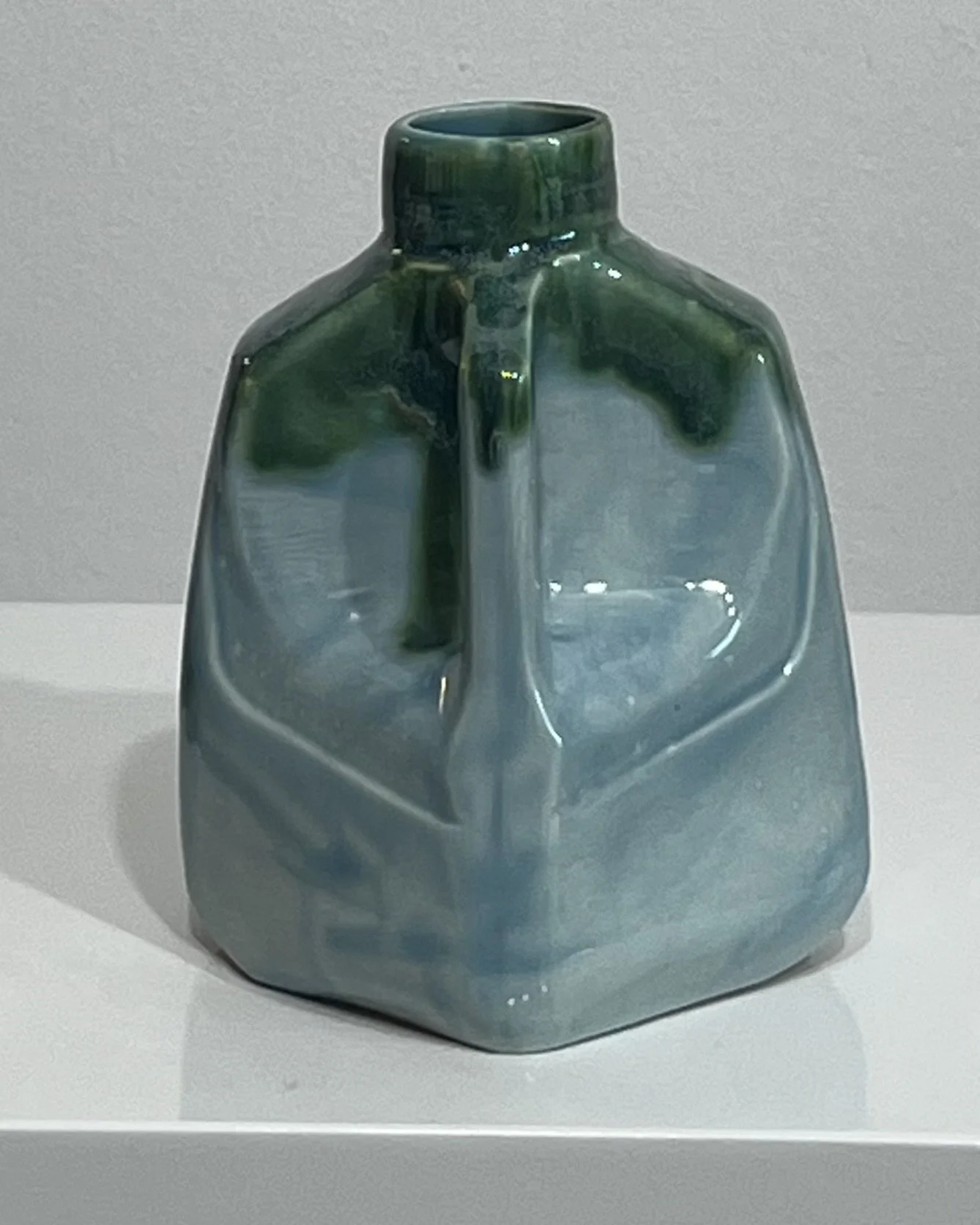 Angled view of blue-green ceramic milk jug sculpture showing its handmade form, handle, and layered glaze.
