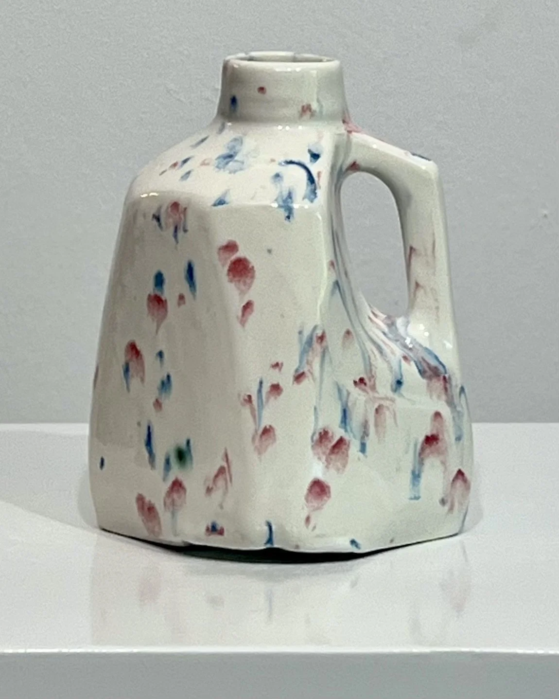 Front view of Kaboom, a handmade porcelain milk jug vase with red and blue splatter glaze on a creamy white surface.