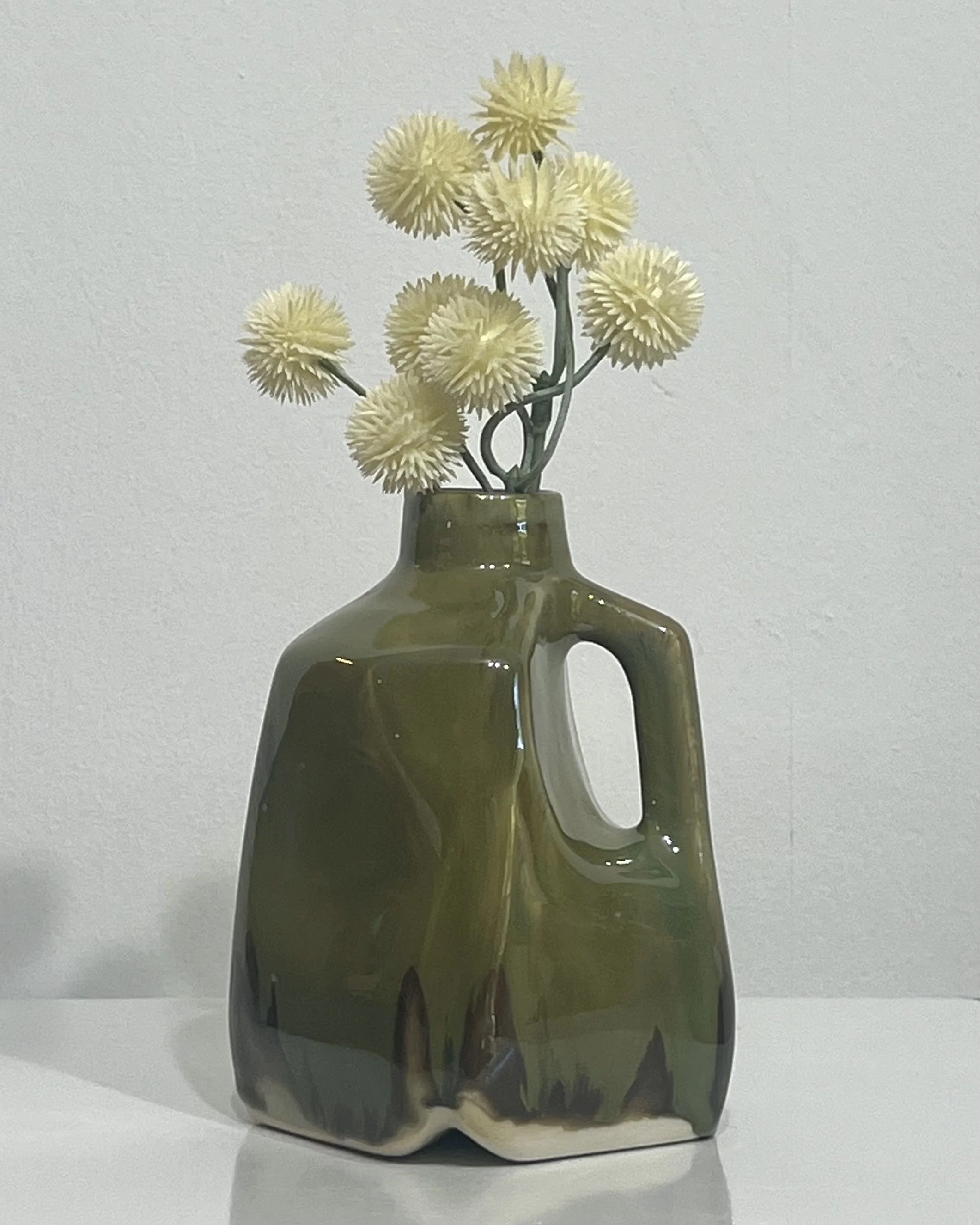 Handmade porcelain milk jug vase by Alissa Van Atta, glazed in olive-green Seaweed and Chun Plum, displayed with yellow dried flowers.