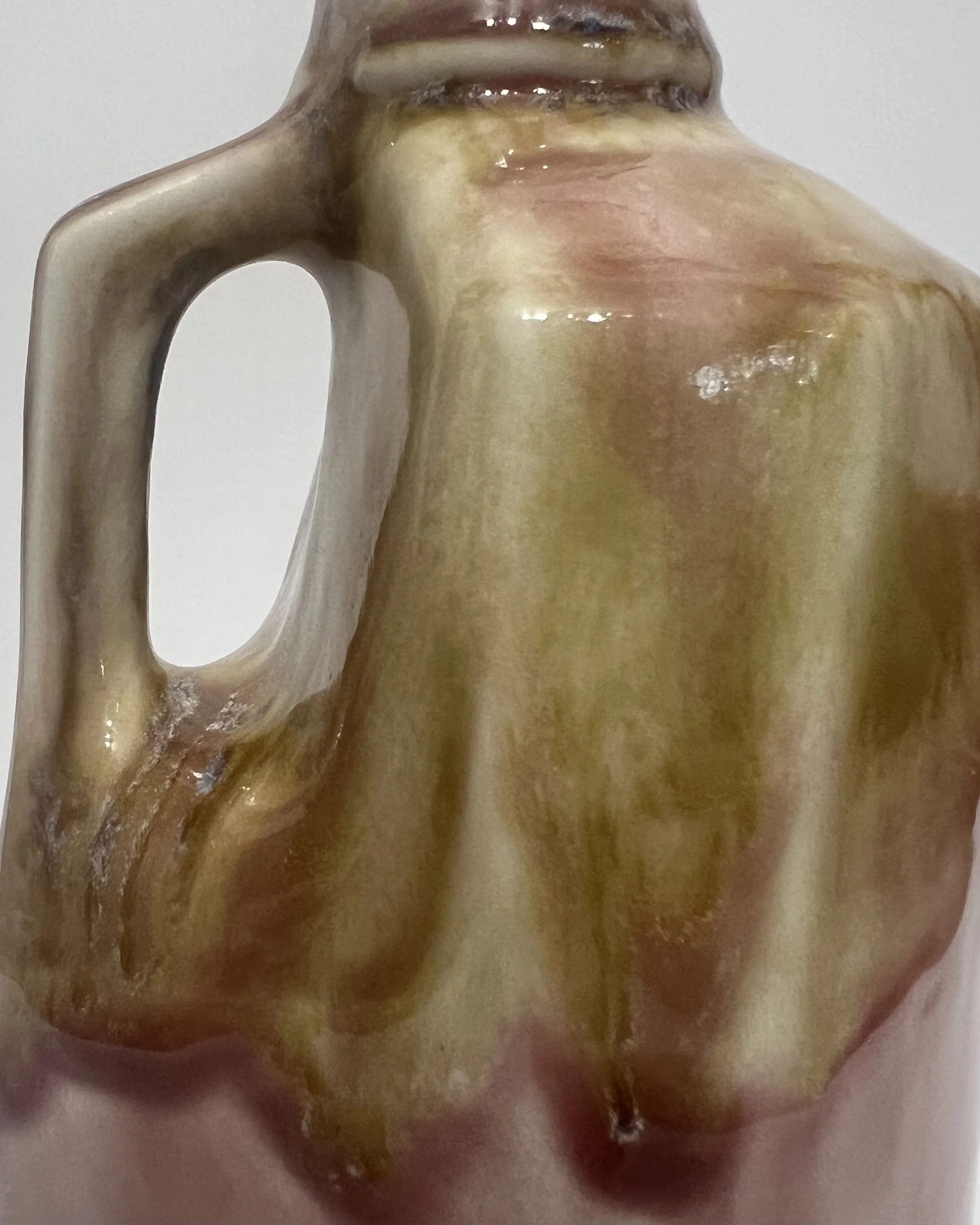 Close-up of tall ceramic milk jug showing layered olive green, blush pink, plum, and cream glaze around the handle.