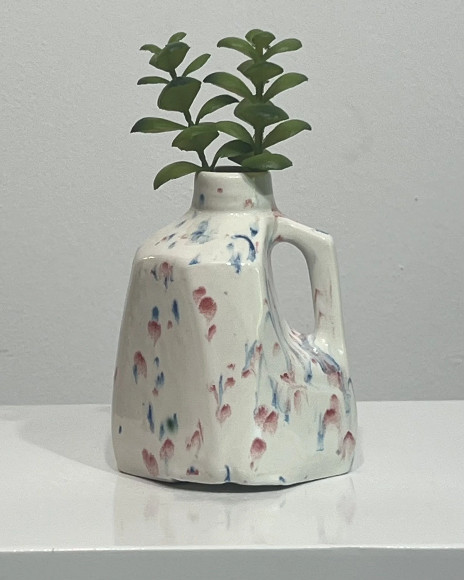 Handmade porcelain milk jug vase with red and blue splatter glaze, displayed with green stems. One-of-a-kind ceramic vase titled Kaboom by North Carolina artist Alissa Van Atta.
