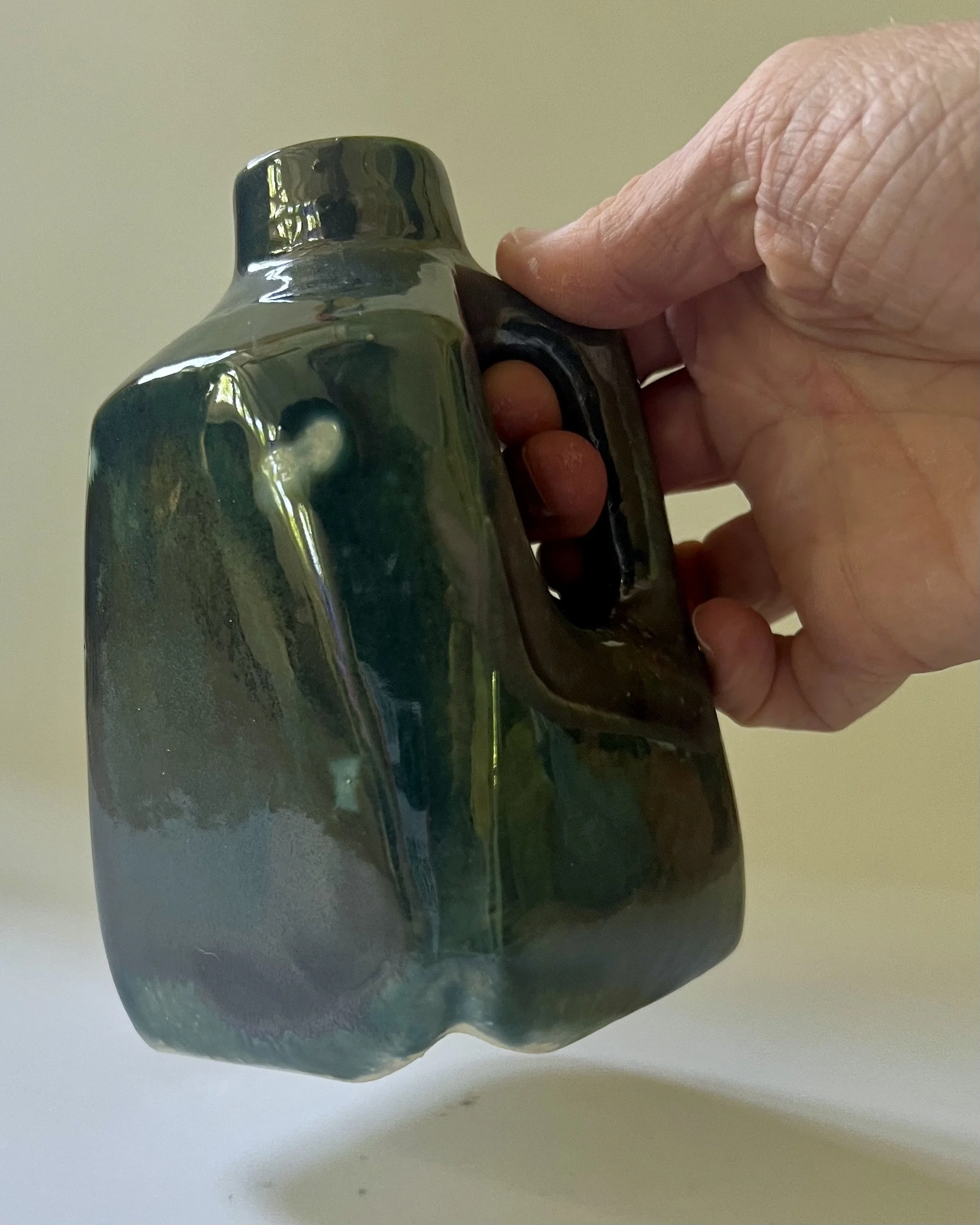 Hand holding handmade porcelain milk jug vase glazed in Vintage Gold over Rainforest, showing scale and sculptural form.