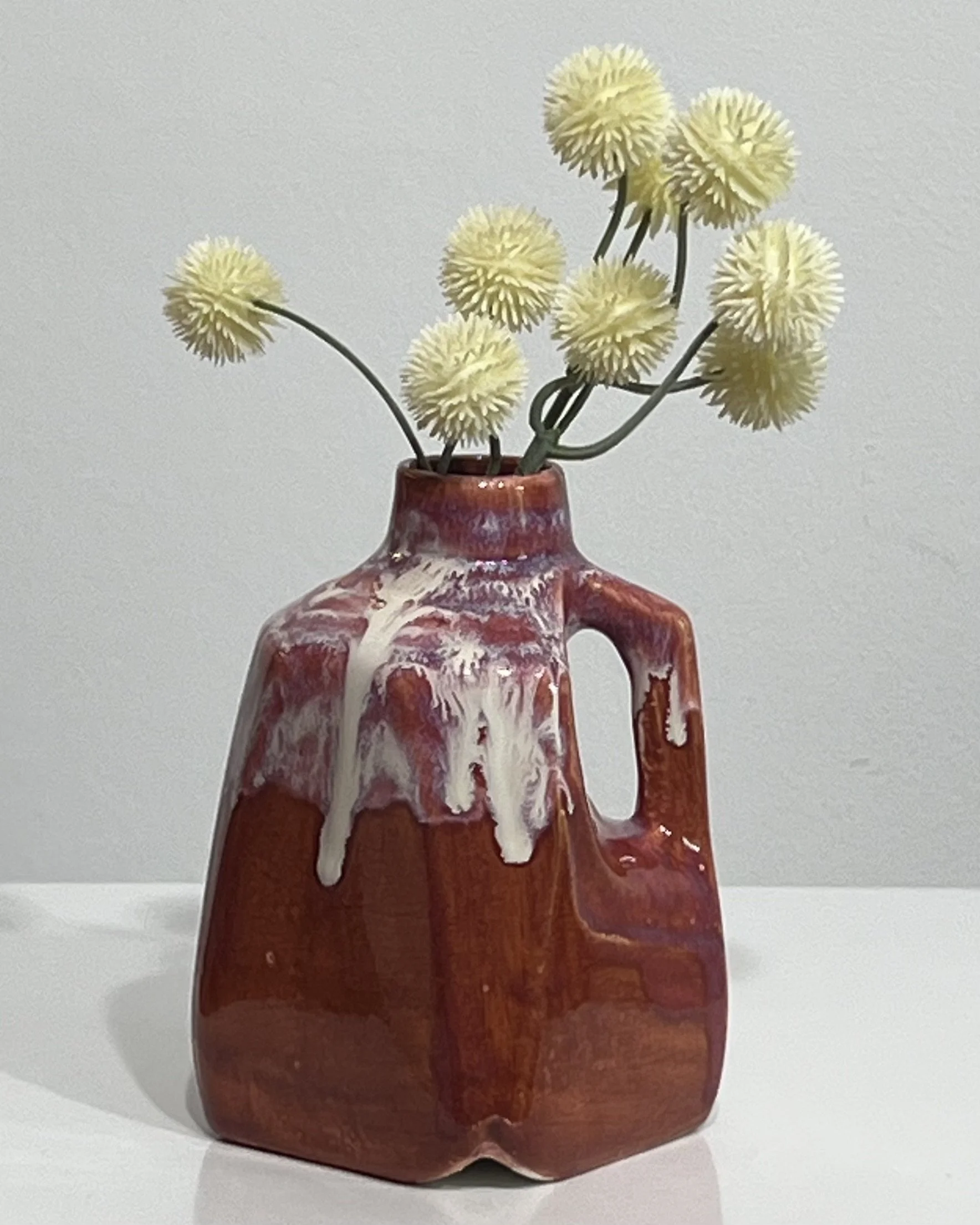 Handmade porcelain milk jug by Alissa Van Atta with deep rust-red Chun Plum and flowing cream Light Flux glazes, displayed with small yellow flowers.