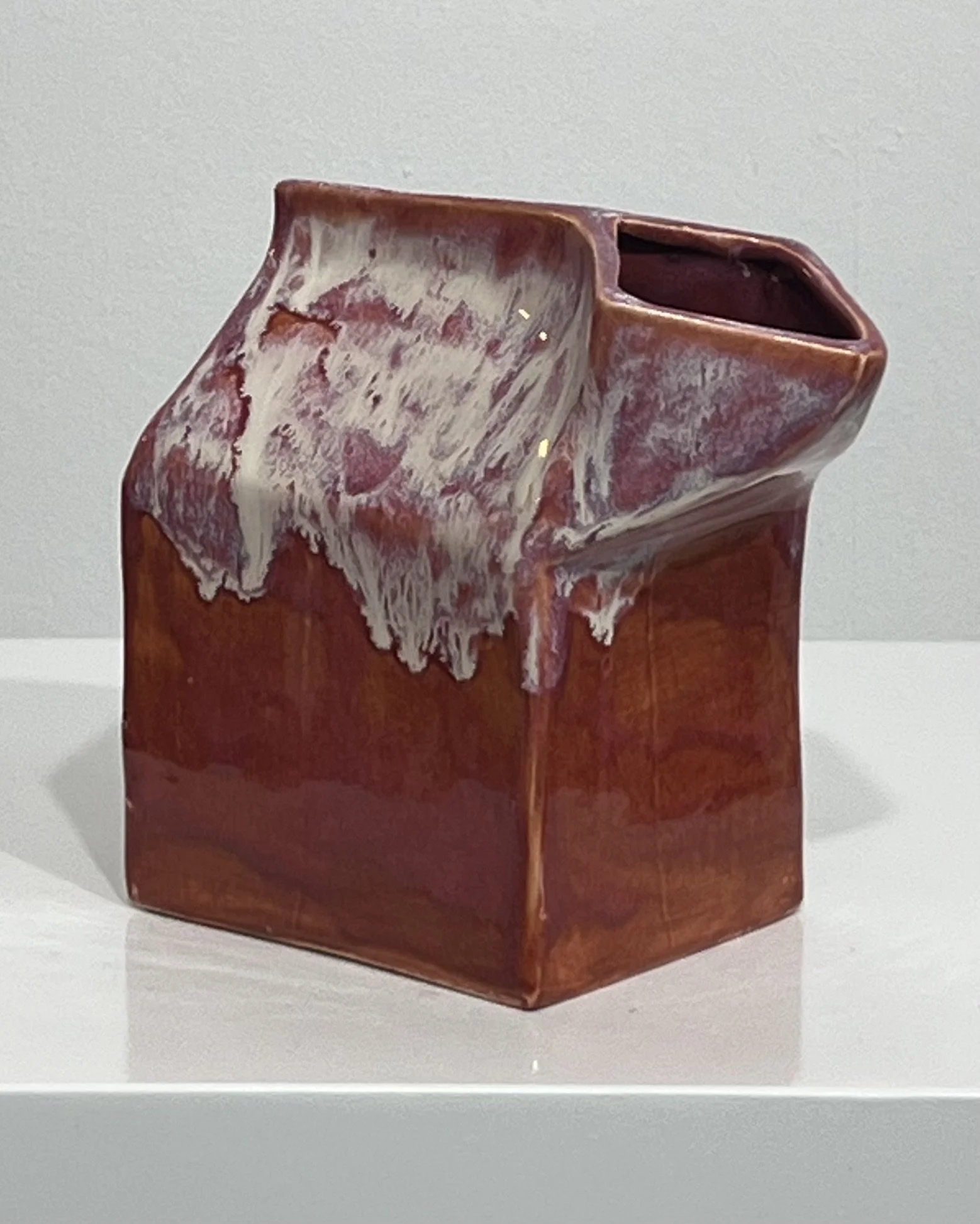 Angled view of the ceramic milk carton showing its asymmetrical folded spout and layered plum and cream glaze.