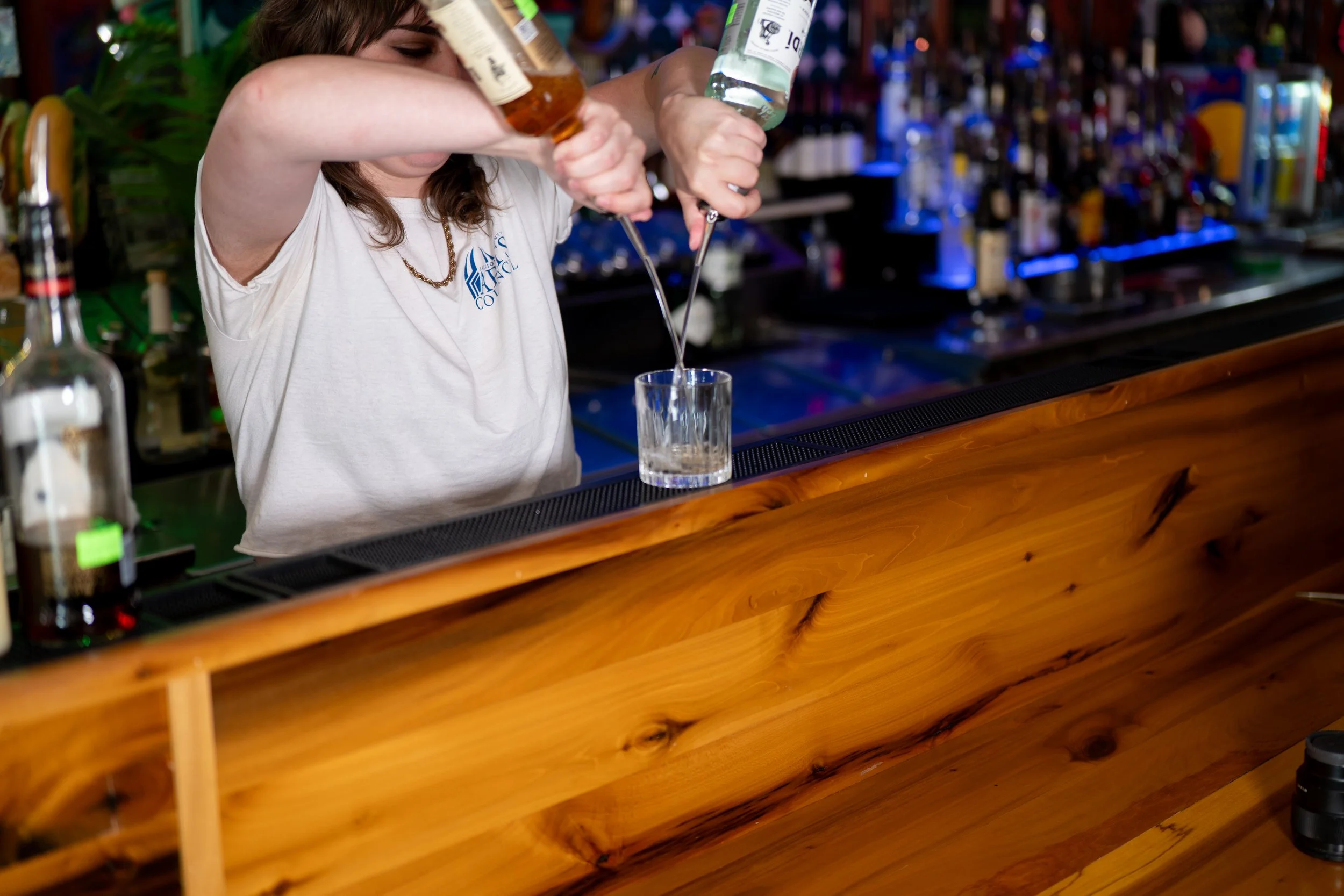 A bartender pouring clear liquid into a glass at a bar with bottles of alcohol in the background.