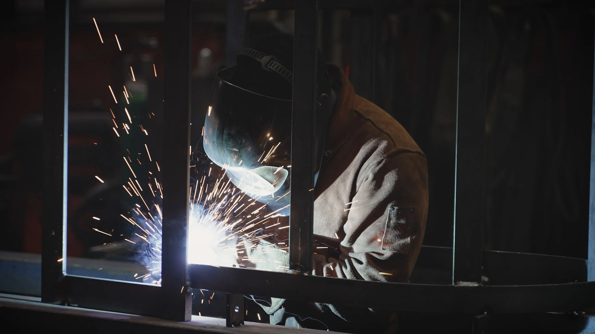 A person welding metal inside a workshop, wearing a protective helmet and brown jacket, with sparks flying from the welding process.