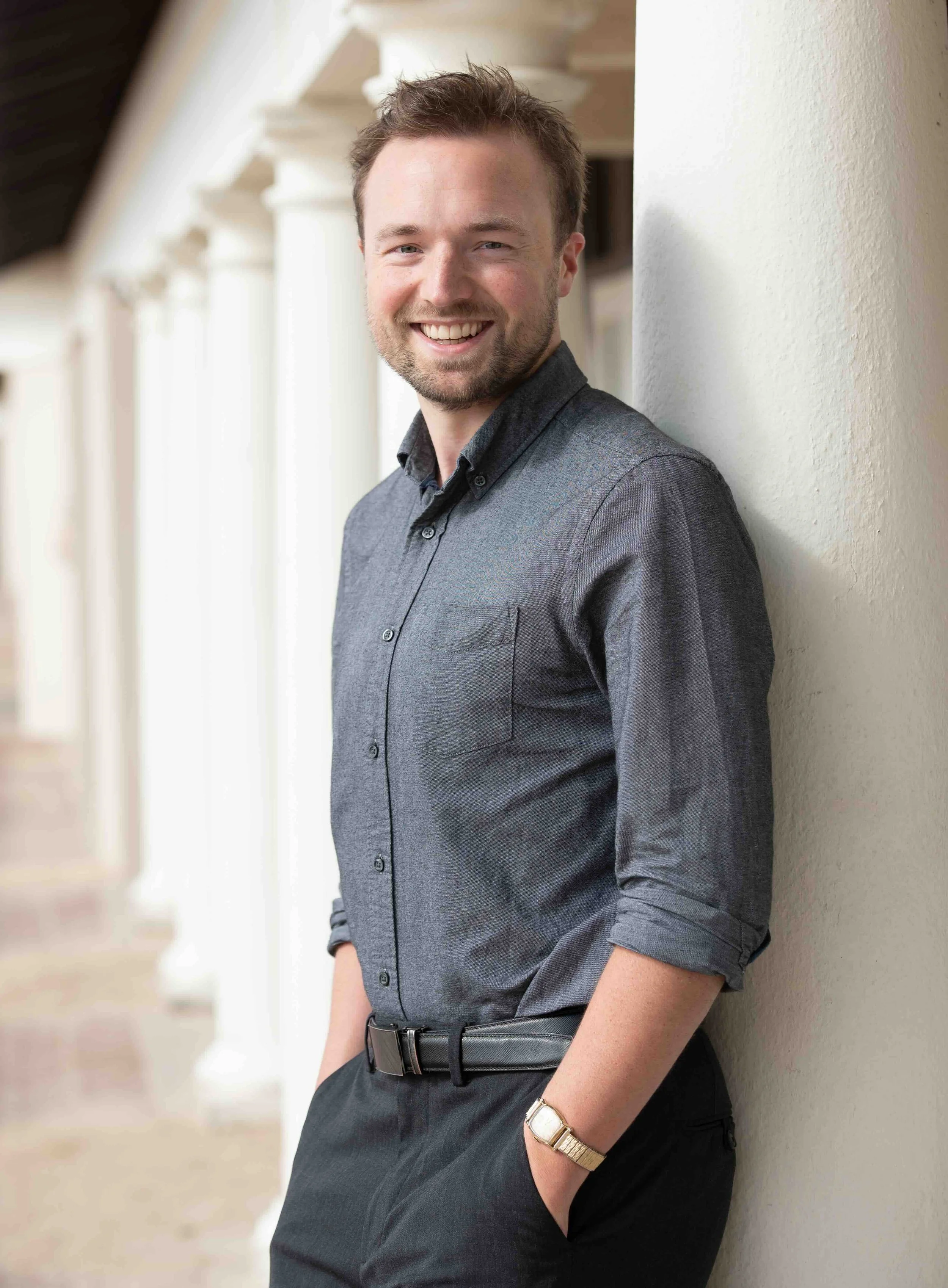 A smiling young man with light brown hair and a beard leaning against a column outdoors. He is wearing a dark gray button-up shirt with rolled-up sleeves, dark pants, and a gold watch.