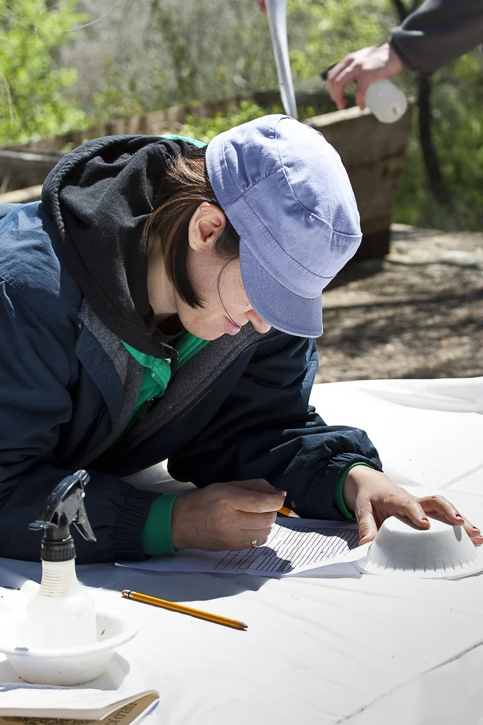 Woman wearing a blue cap and jacket writing on a sheet of paper outdoors, surrounded by trees, with a person in the background holding a container.