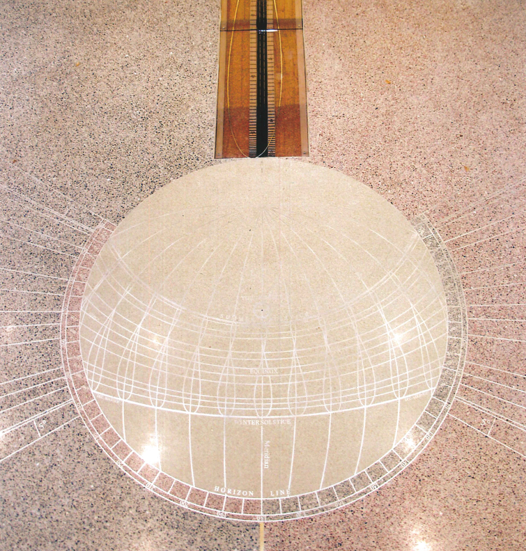 A glass globe-shaped skymap sundial with grid lines and labels, embedded in a speckled sidewalk, and a metal gnomon casting a shadow to tell time.