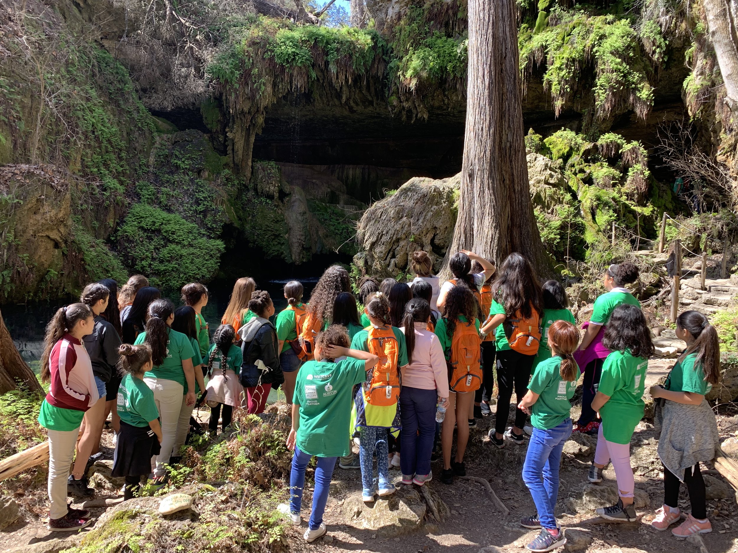 A large group of people, many young, gathered outdoors near a rocky and green cave or grotto, with some trees and sunlight overhead.