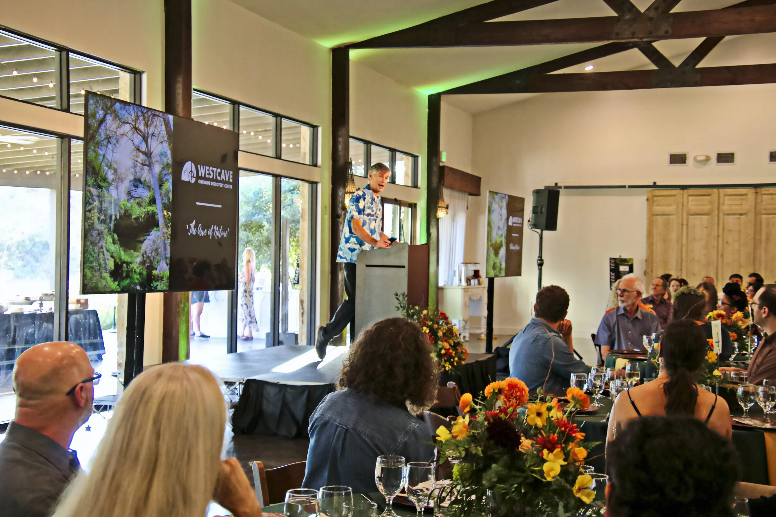 A man standing at a podium speaking to an audience at an indoor event, with a large screen displaying the logo for Westcave Outdoor Discovery Center and an outdoor scene with trees in the background.