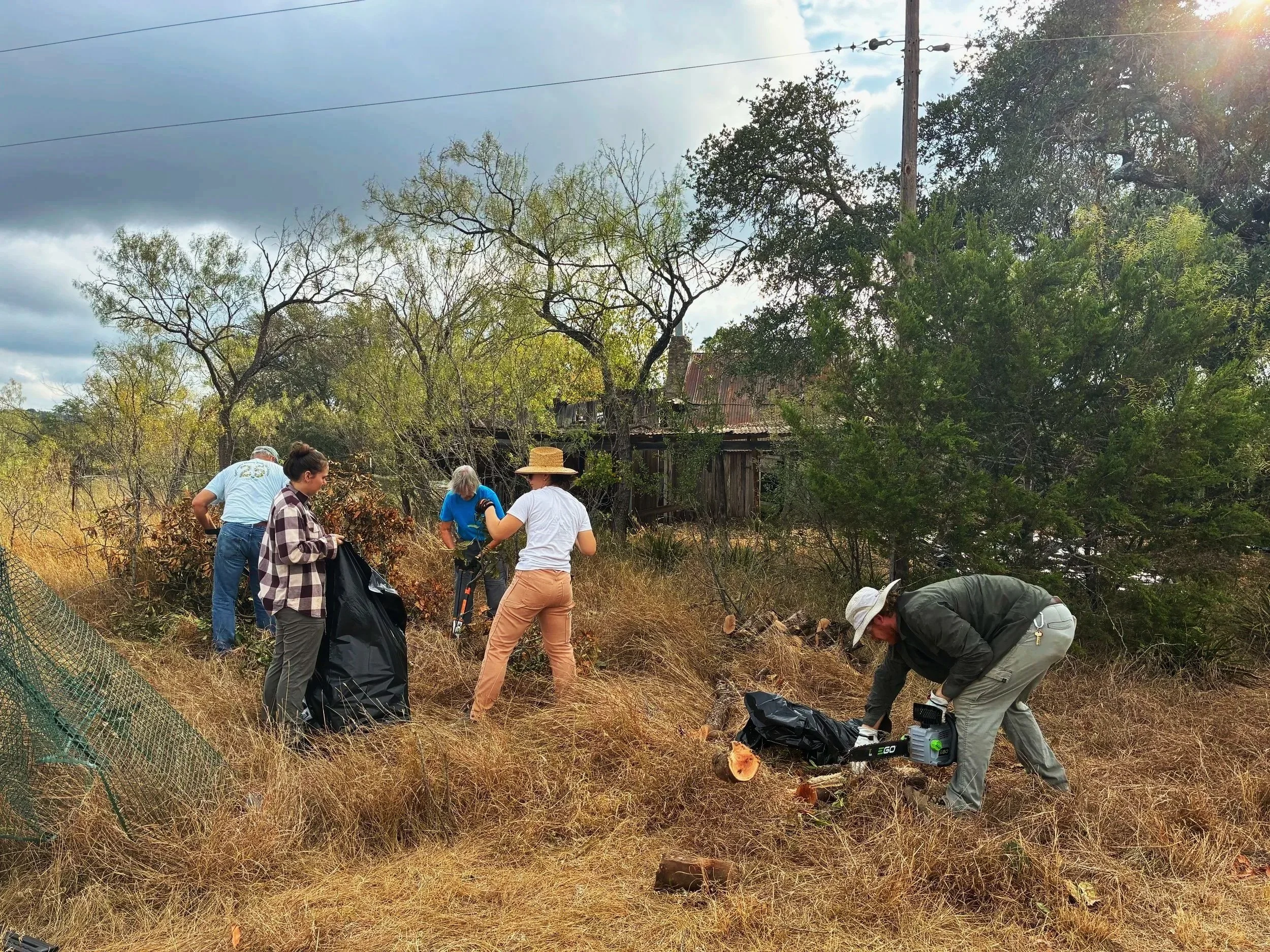 A group of people working outdoors in a rural area, with trees and an old wooden house in the background, some using tools and wearing hats.