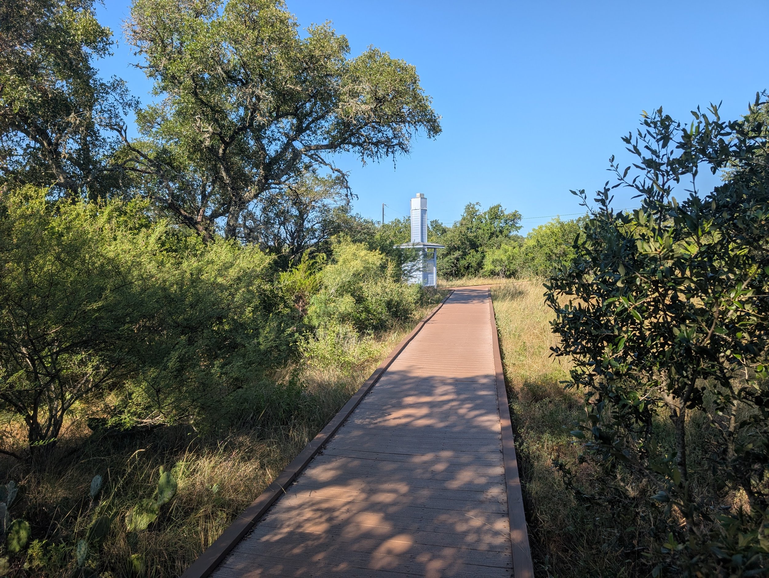 A wooden path running through a lush green area lined with trees and bushes, with a tall chimney swift tower visible in the background under a clear blue sky.